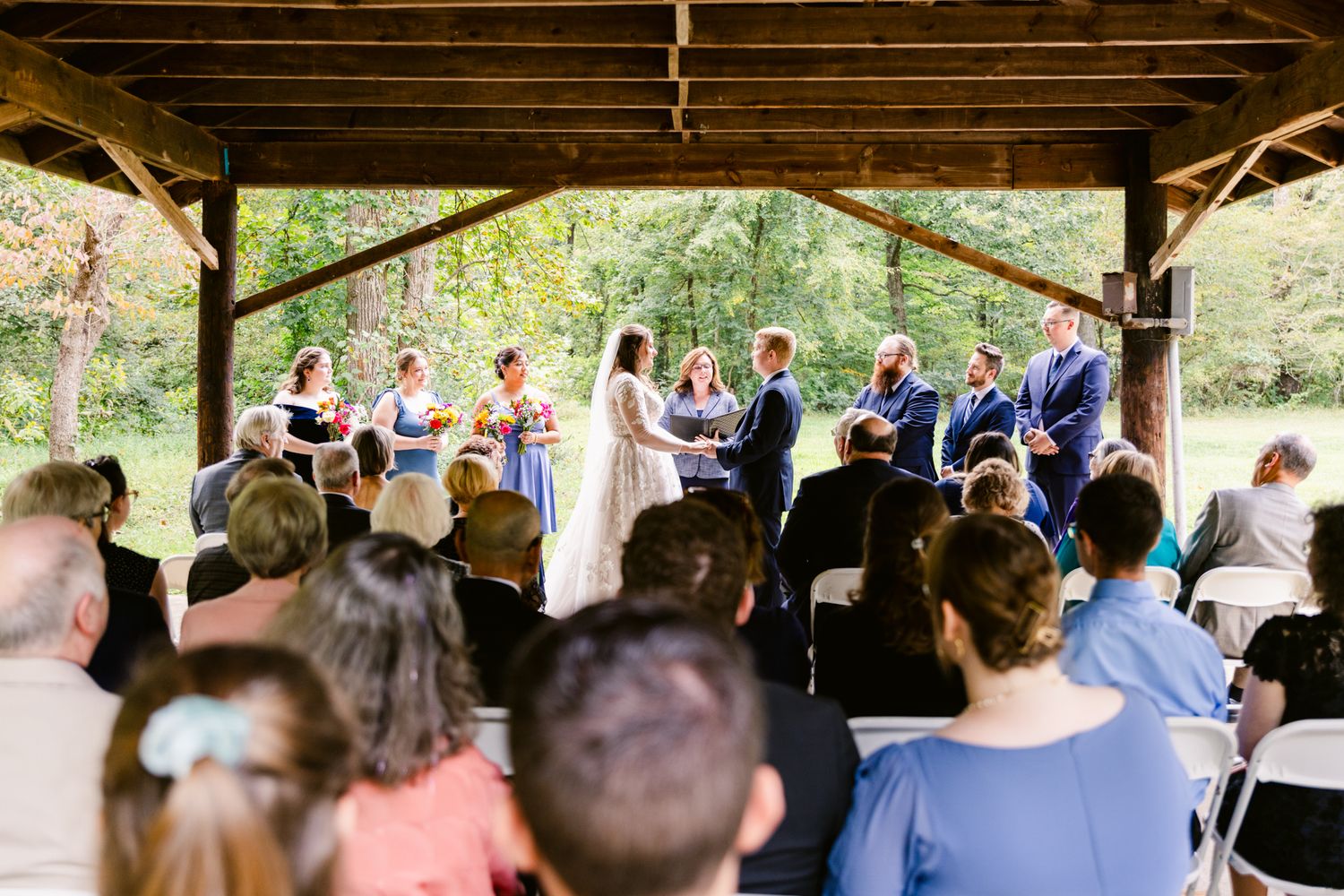 Wedding guests gather under a rustic wooden pavilion for an outdoor ceremony.
