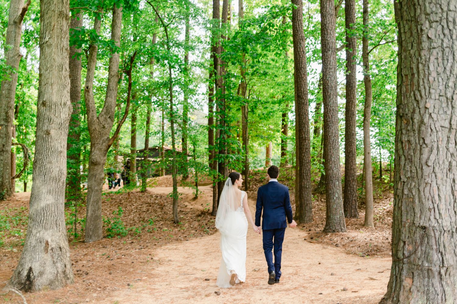 Newlyweds walk hand in hand through a serene forest path lined with tall trees.