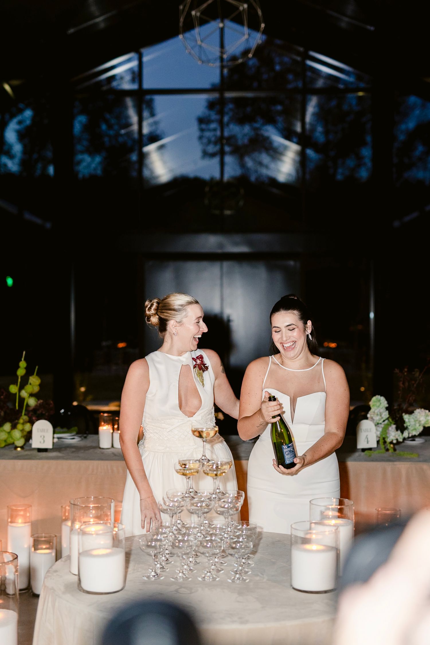 Wedding party members in white attire laugh together at a candlelit reception table with champagne glasses.
