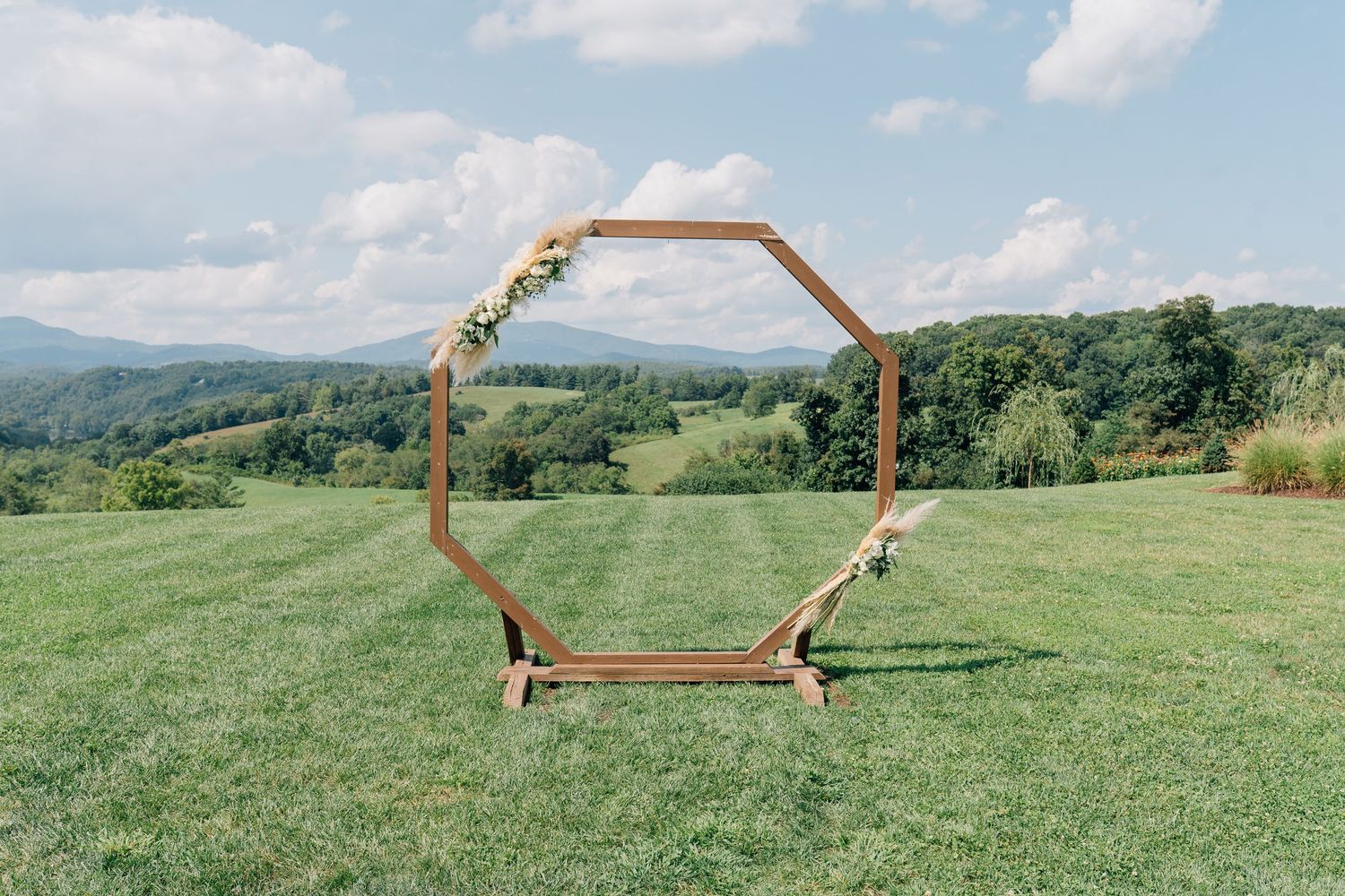 A wooden arch decorated with flowers stands in a green meadow with rolling hills and mountains in the background.