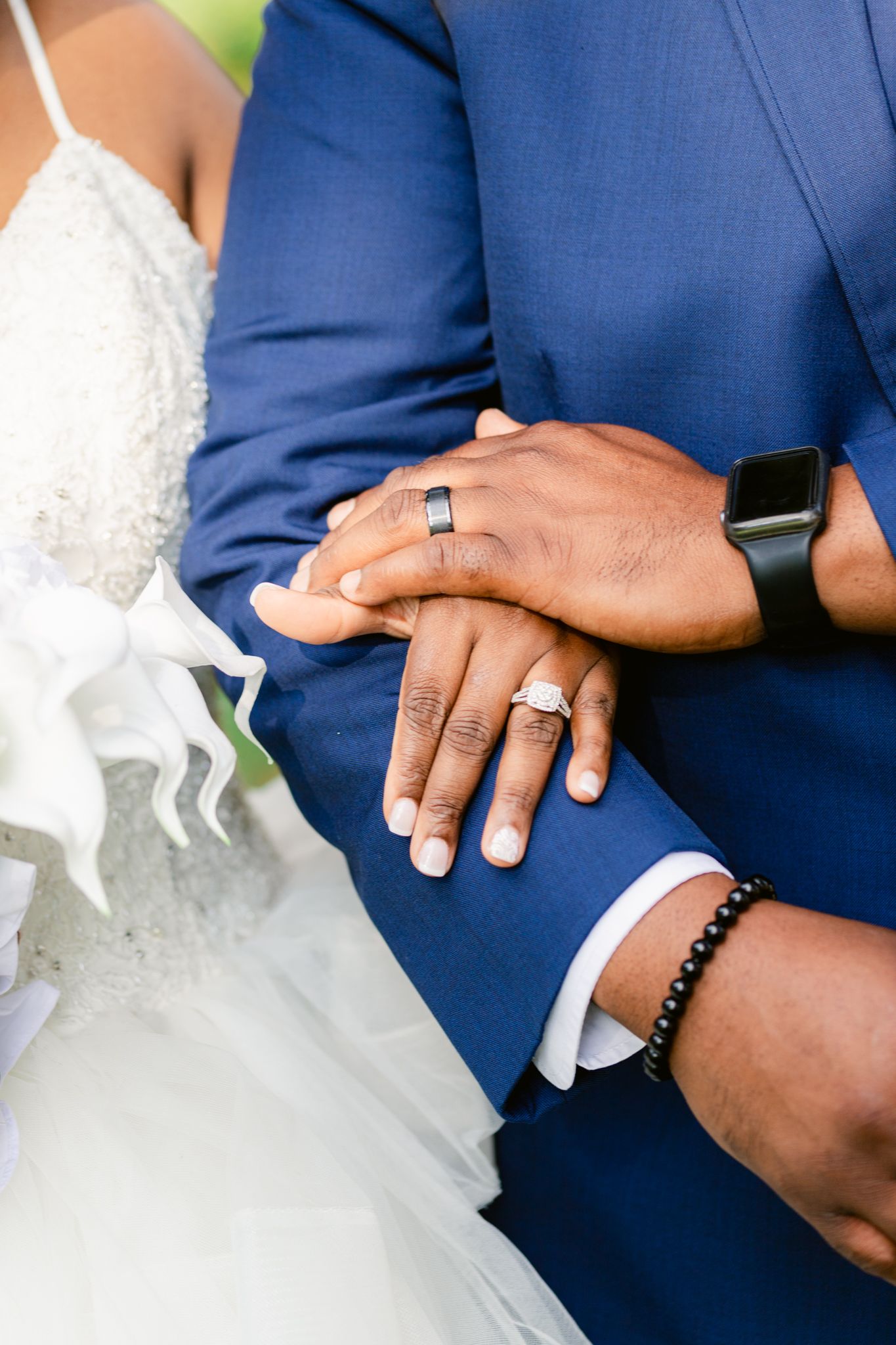 Close-up view of couple's hands showing wedding rings and formal attire with blue suit and white dress details.