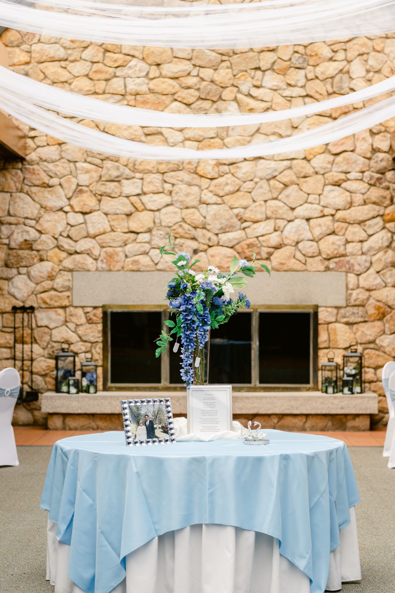 Round tables with light blue tablecloths and white overlays set against a rustic stone wall feature elegant floral centerpieces.
