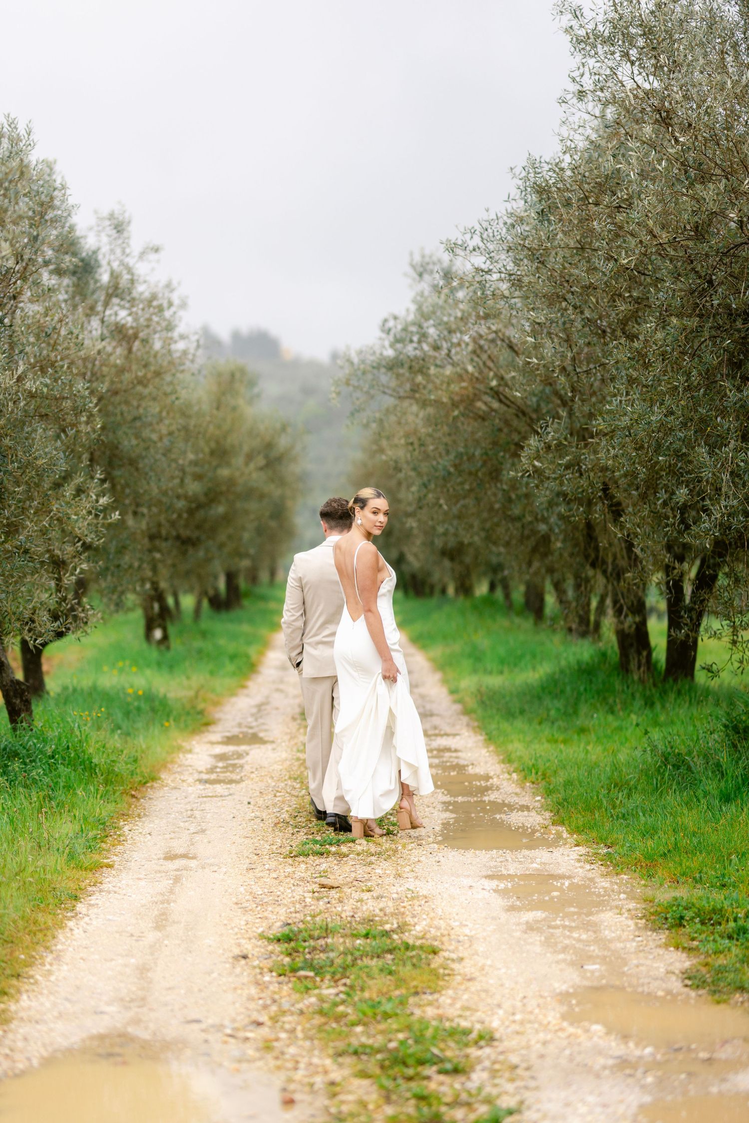 A figure in a white dress walks along a dirt path through an olive grove on a misty day.
