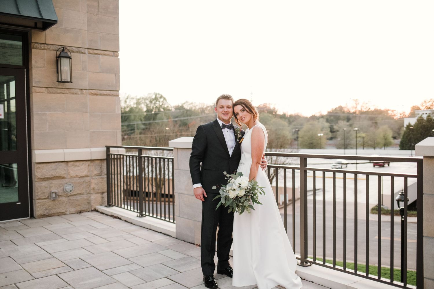 A couple shares a romantic moment on an elegant stone balcony overlooking a scenic landscape at sunset.