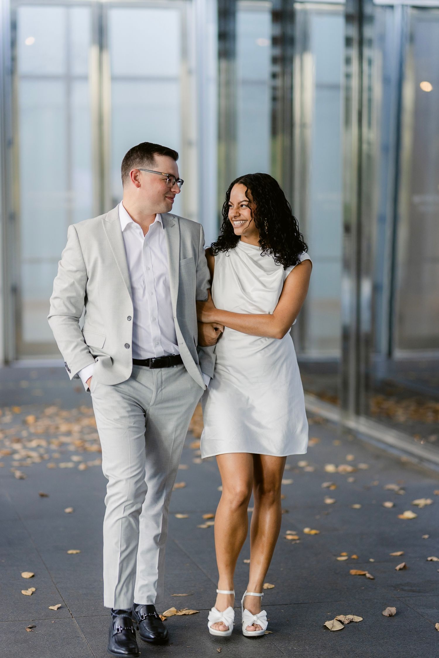 A couple in white outfits poses playfully in a modern glass corridor with fallen autumn leaves on the ground.