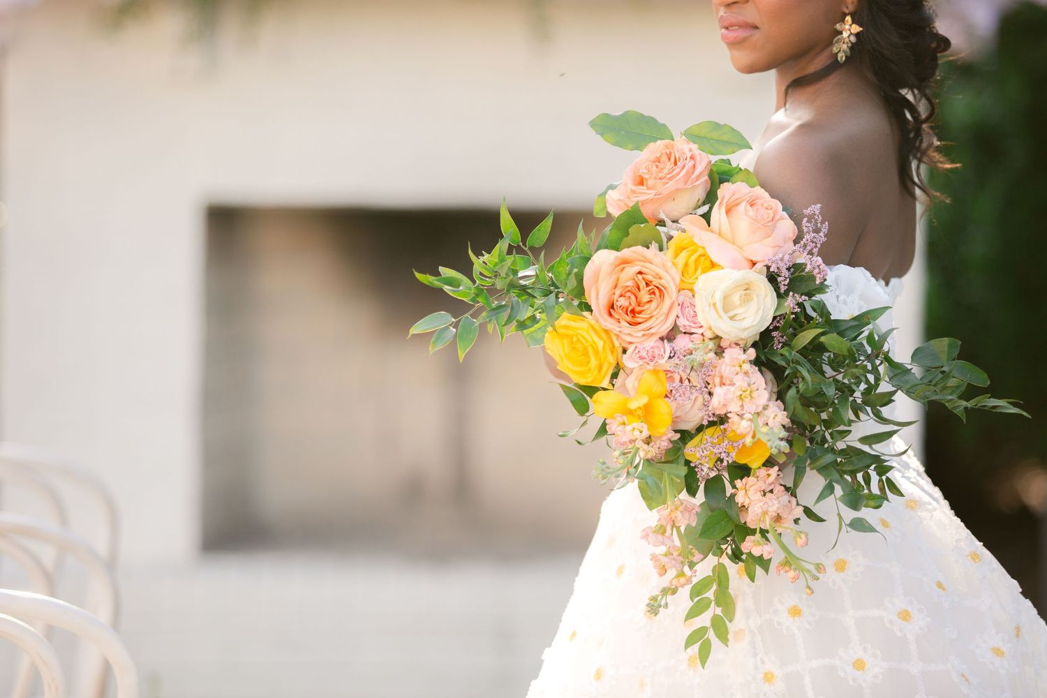 A beautiful bridal bouquet featuring peach, yellow and pink roses with lush greenery against a white wedding dress.