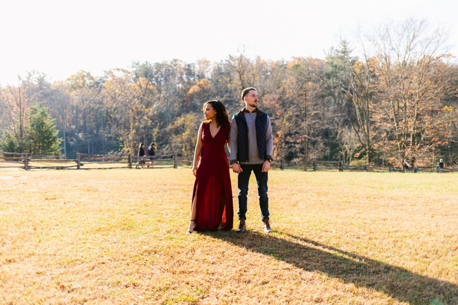 A couple stands together in a golden autumn field with trees in the background on a sunny afternoon.