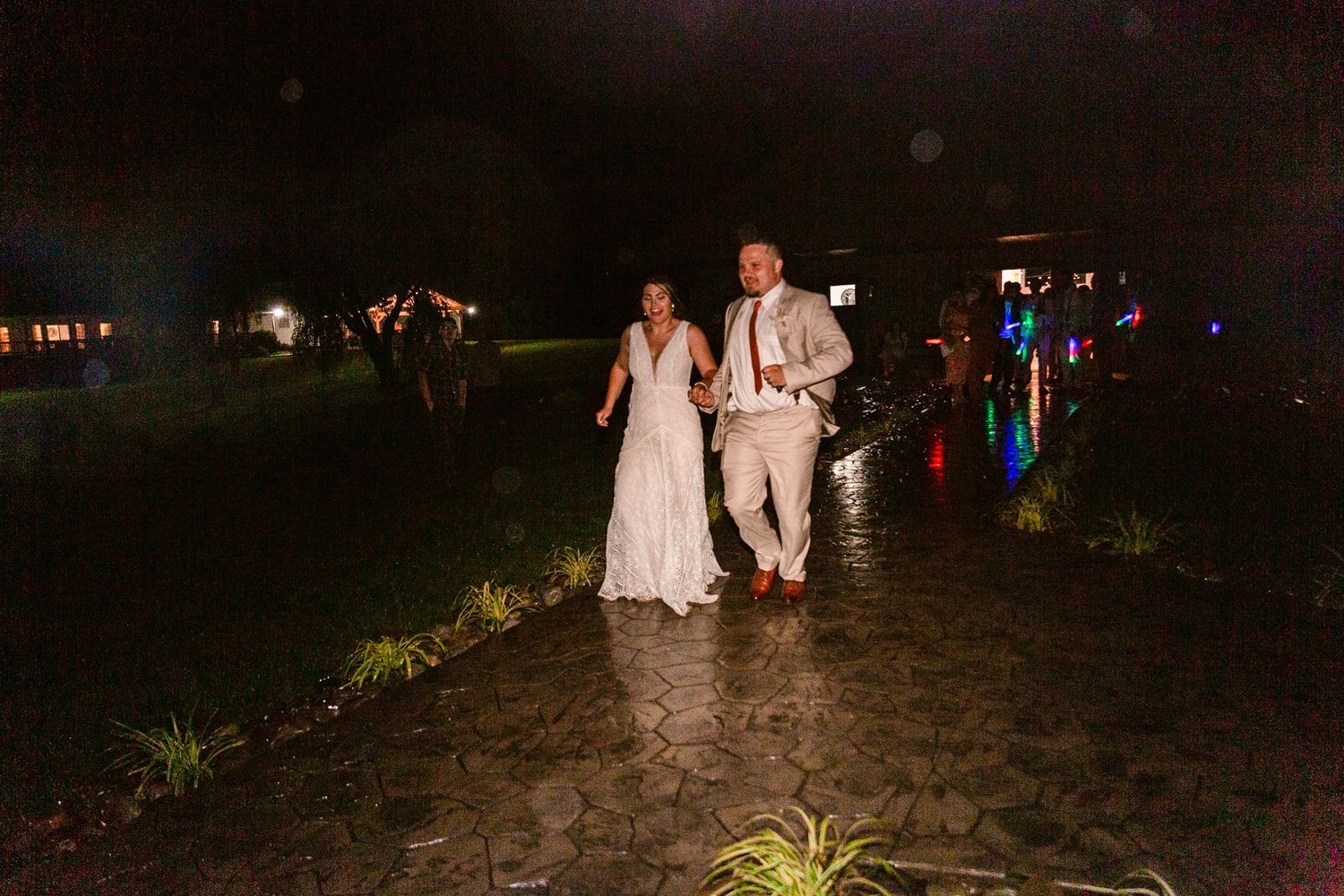 Wedding couple walks along a dimly lit beach path at night with guests following behind them.