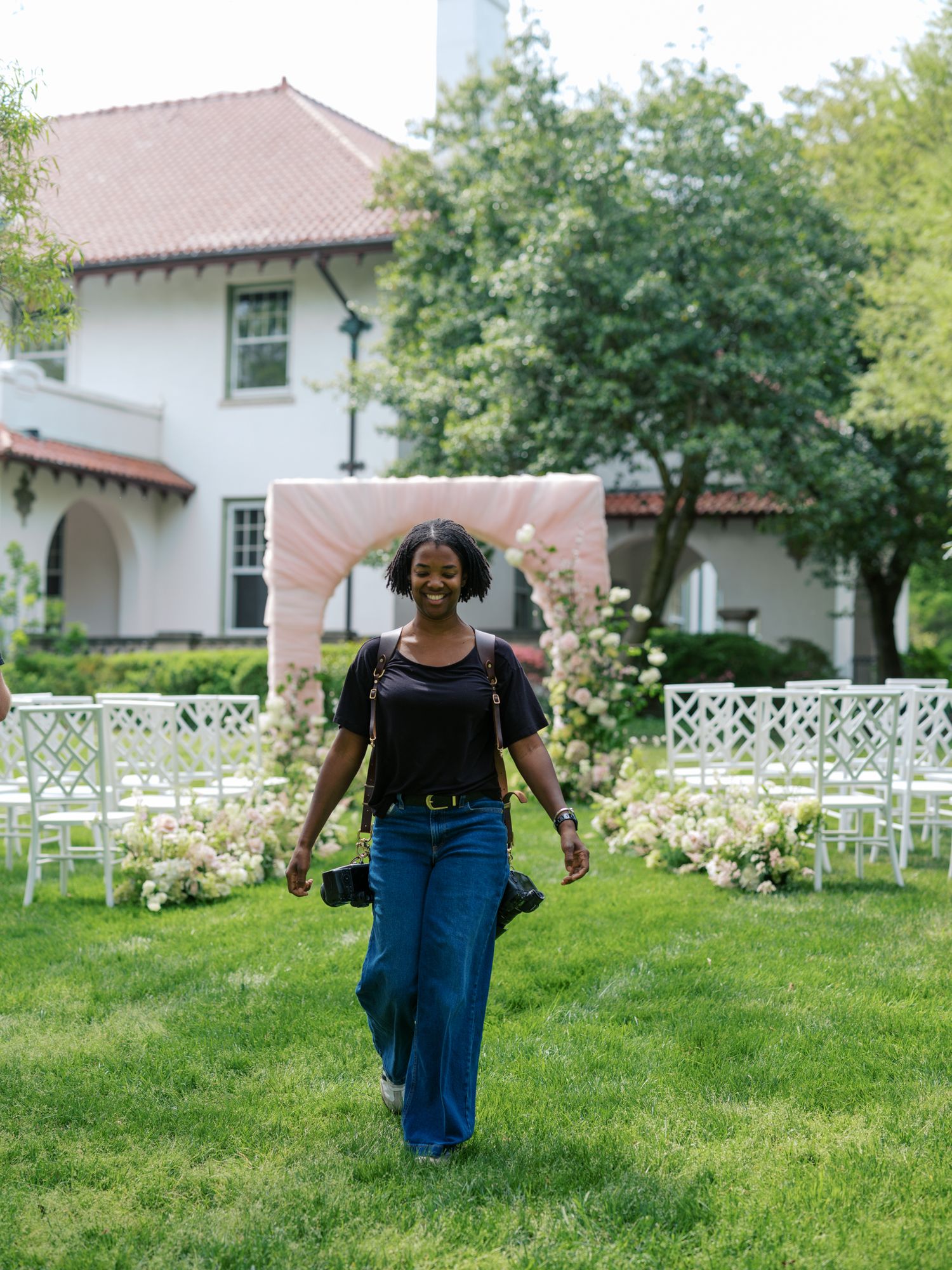 A smiling woman in a black top and wide-leg jeans walks through an outdoor wedding venue with floral arch and white chairs.