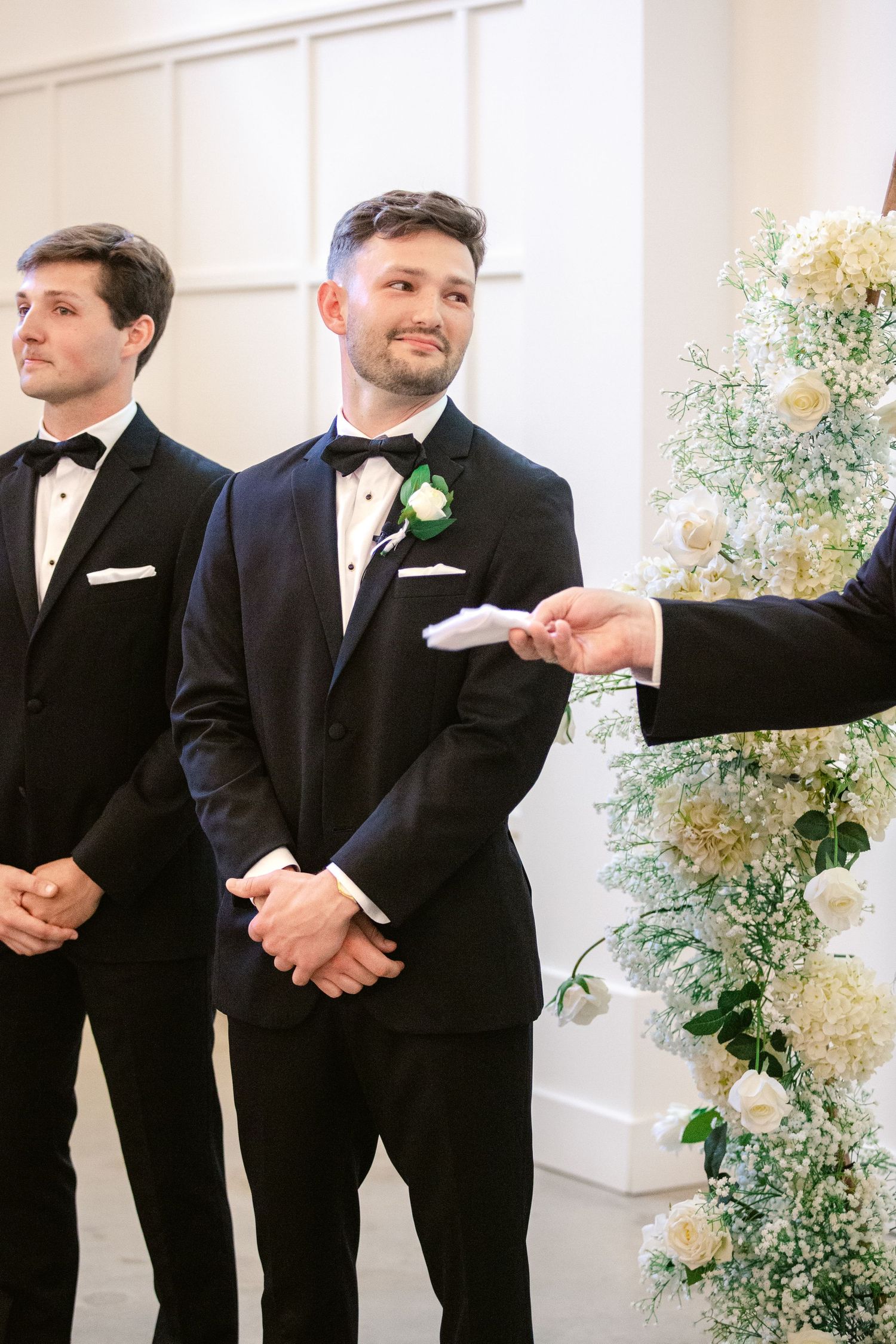 Wedding ceremony with floral arch and groomsmen in black tuxedos at an elegant white venue.