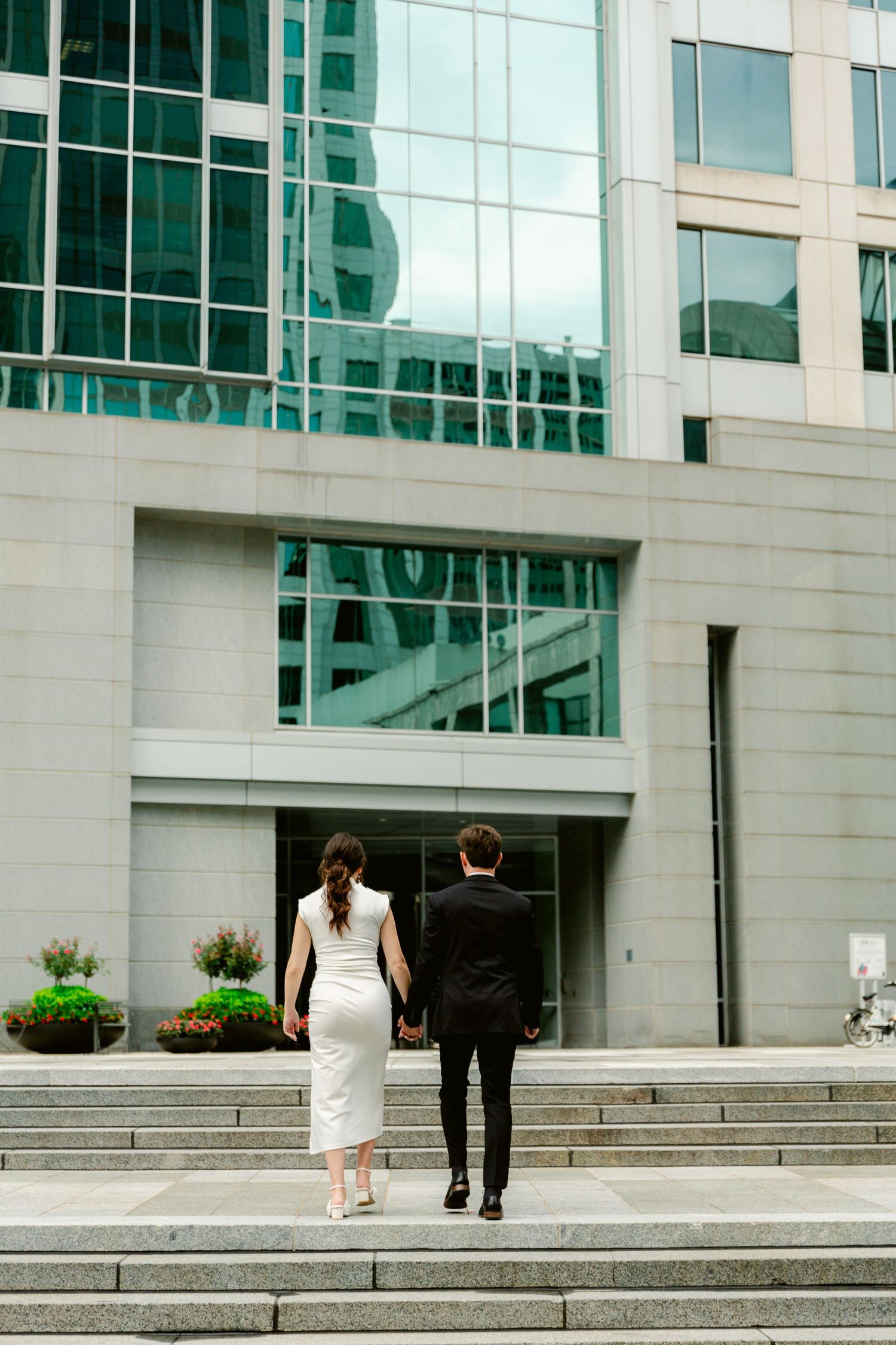 Couple in formal attire walking up concrete steps toward modern glass office building entrance.