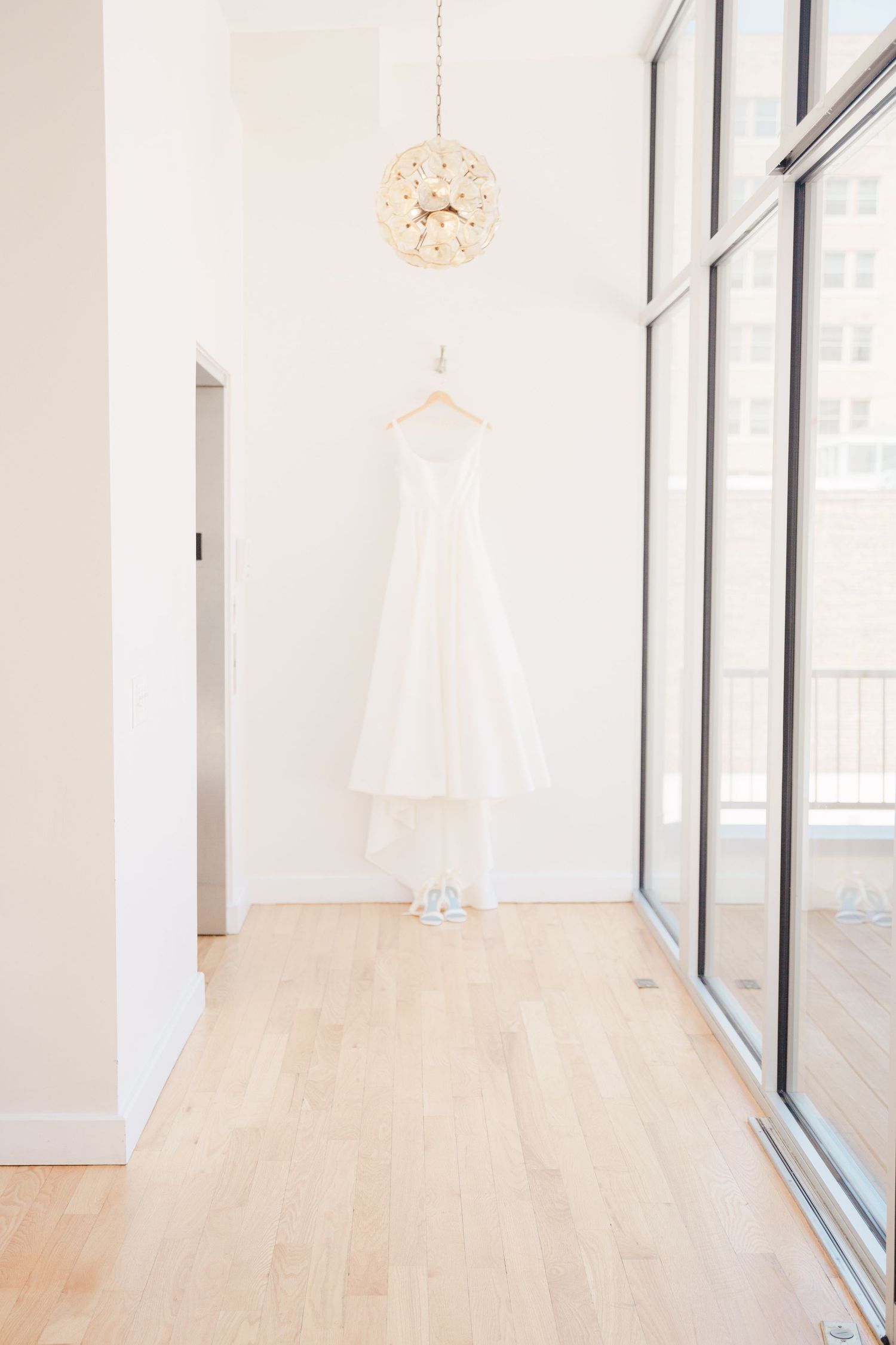 A white wedding dress hangs elegantly in a bright minimalist room with floor-to-ceiling windows and light wood flooring.