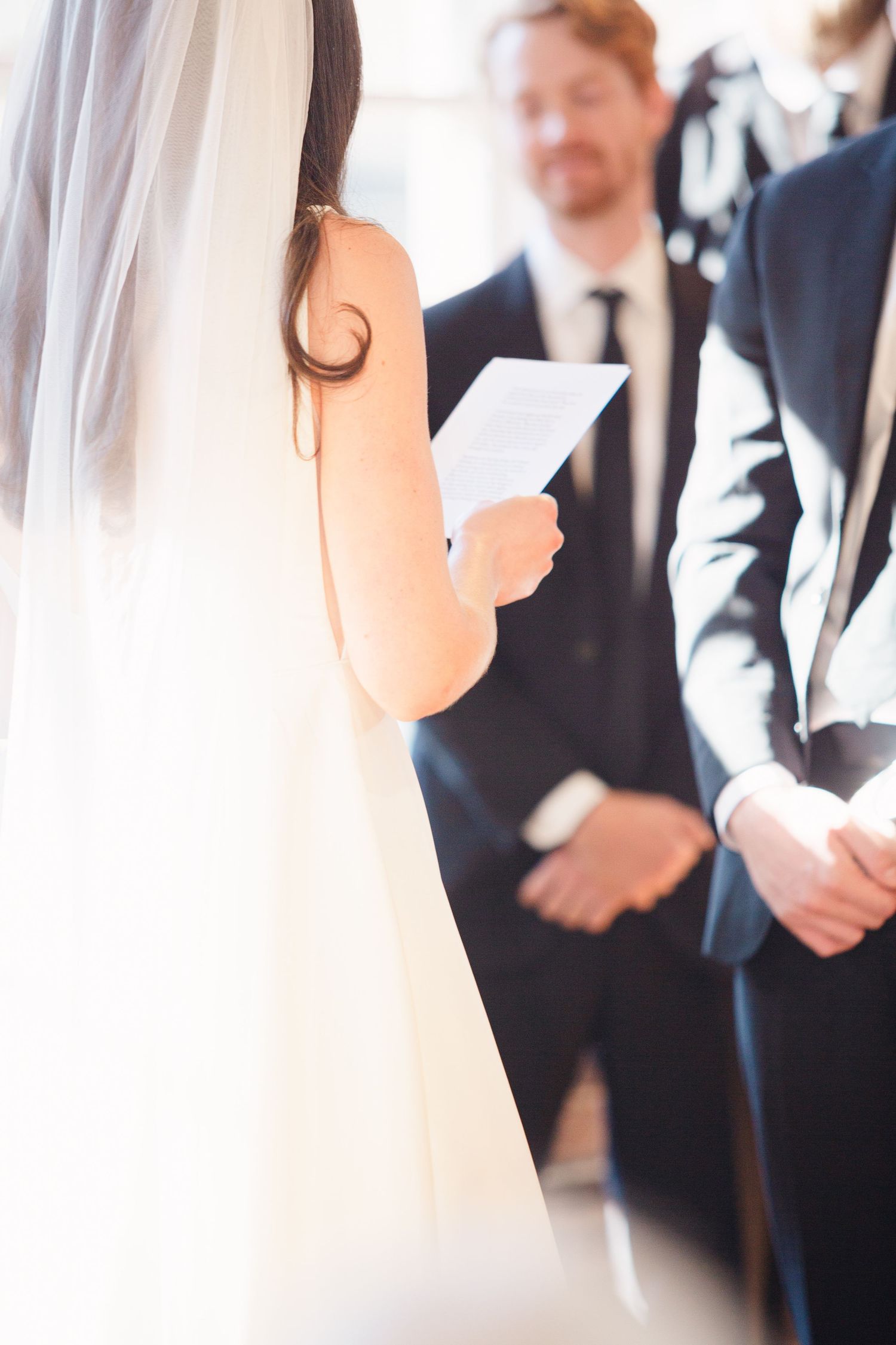 Close-up shot of a bride in white dress reading from paper during wedding ceremony.