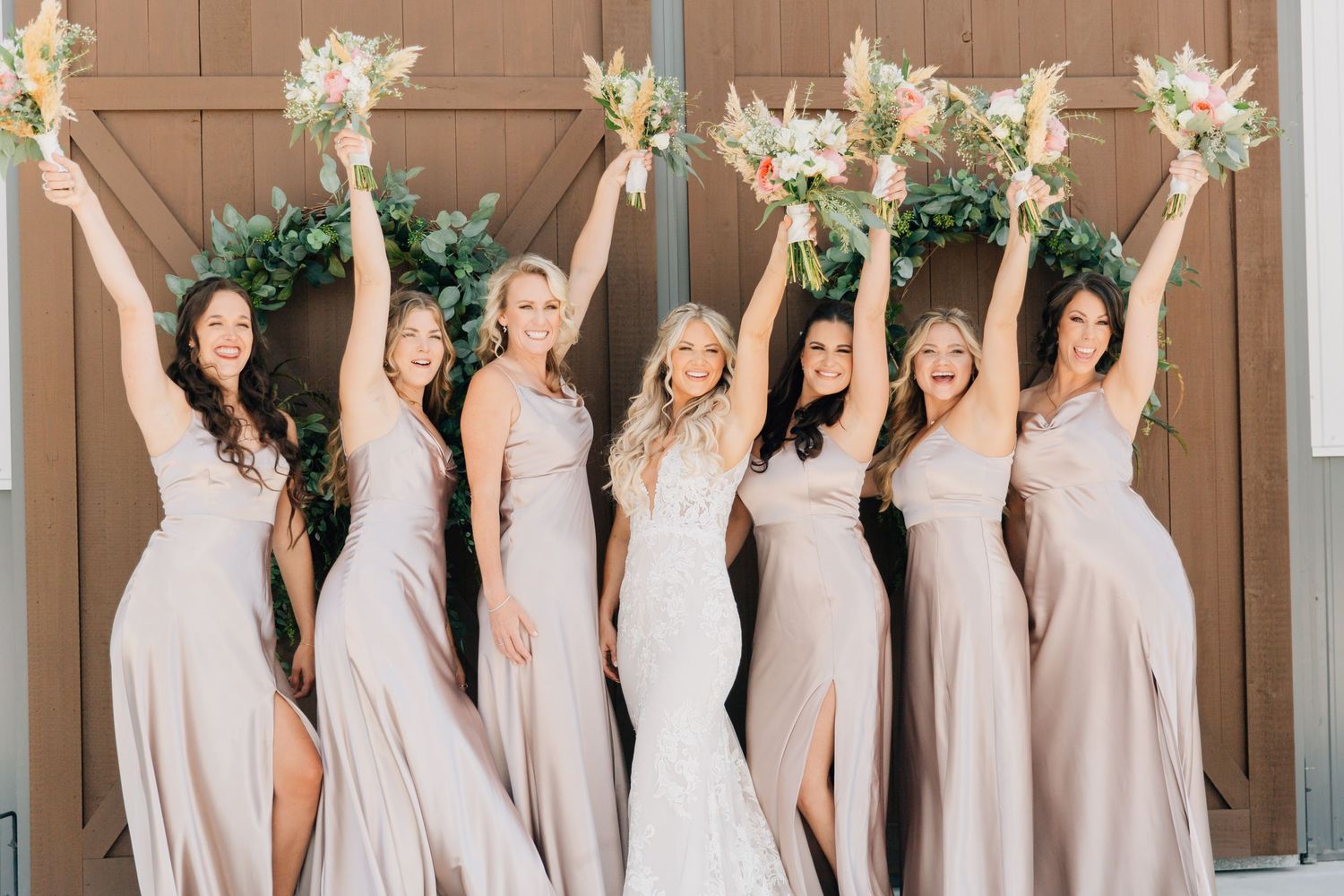 Bridesmaids in champagne dresses holding white bouquets pose joyfully against brown doors.