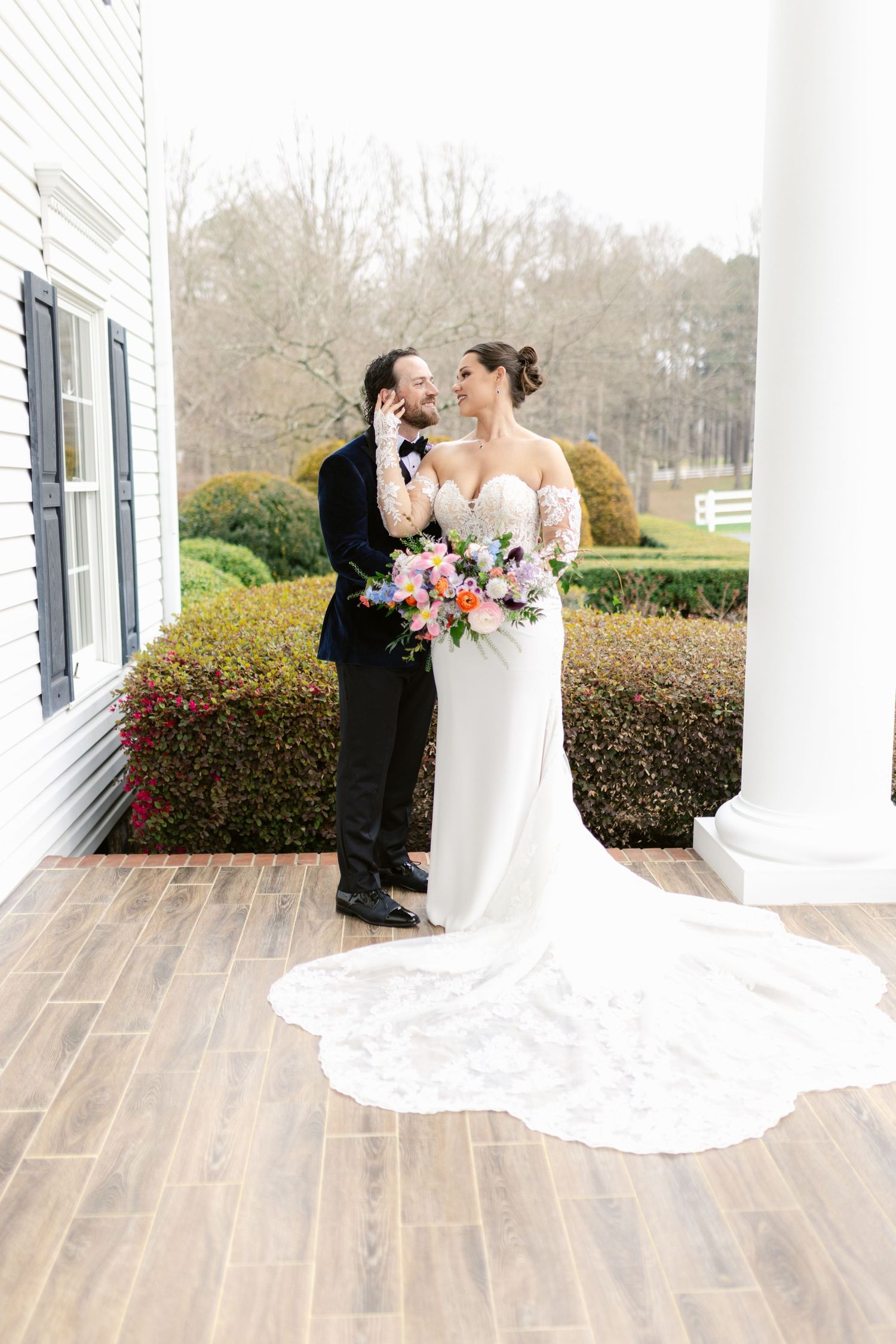A romantic wedding moment on a porch with white columns and evergreen landscaping, featuring an elegant long-train dress.