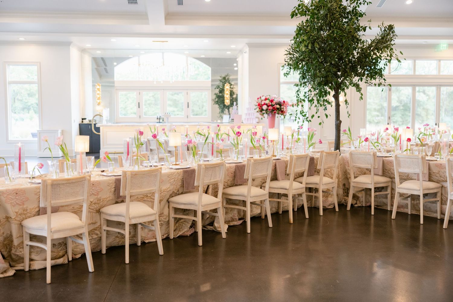 Long banquet tables with white linens and floral centerpieces set up in an elegant reception hall with large windows.