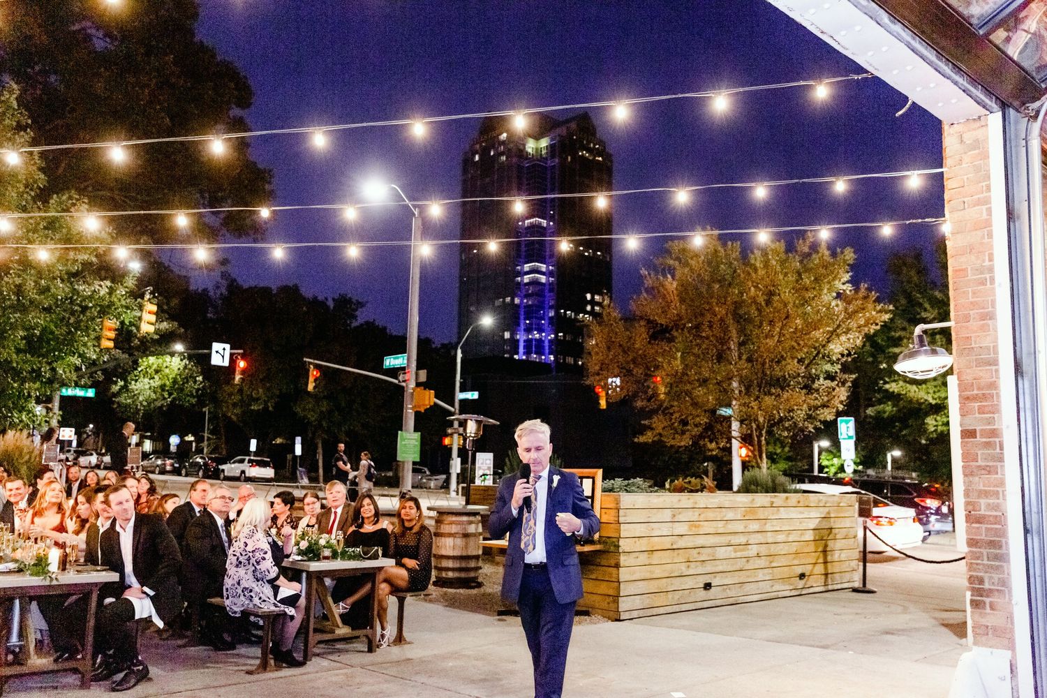 String lights illuminate an outdoor evening venue with trees and guests mingling under a covered patio area.