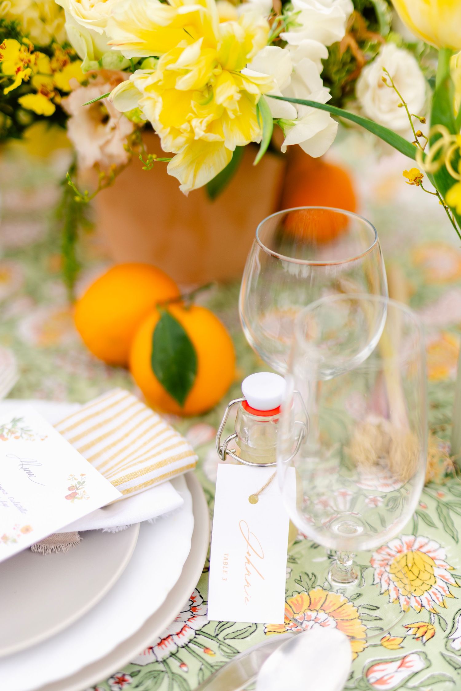 A spring table setting with daffodils, oranges, wine glasses and floral tablecloth in soft natural light.
