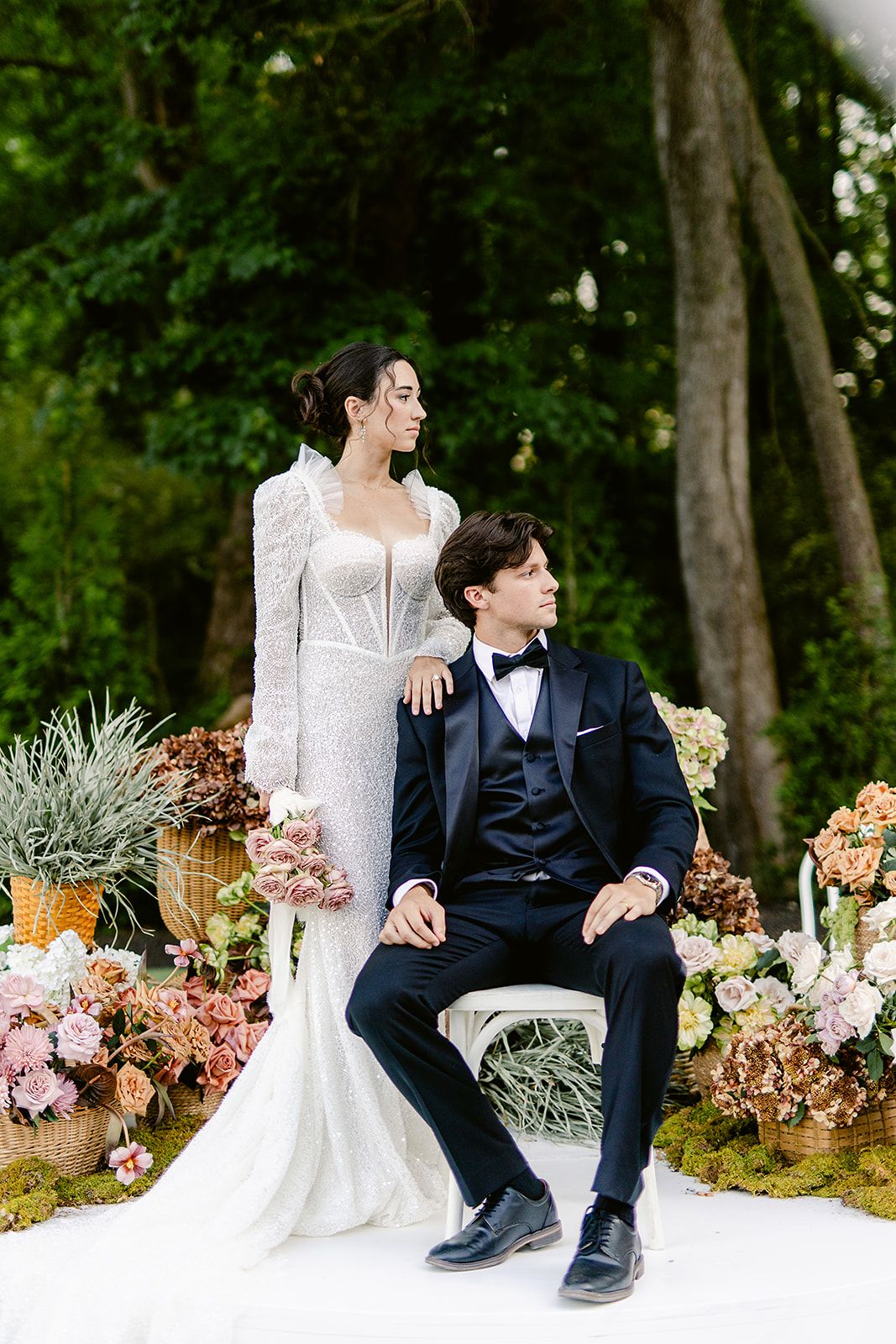 A couple in wedding attire sits on a bench surrounded by lush greenery and colorful floral arrangements.