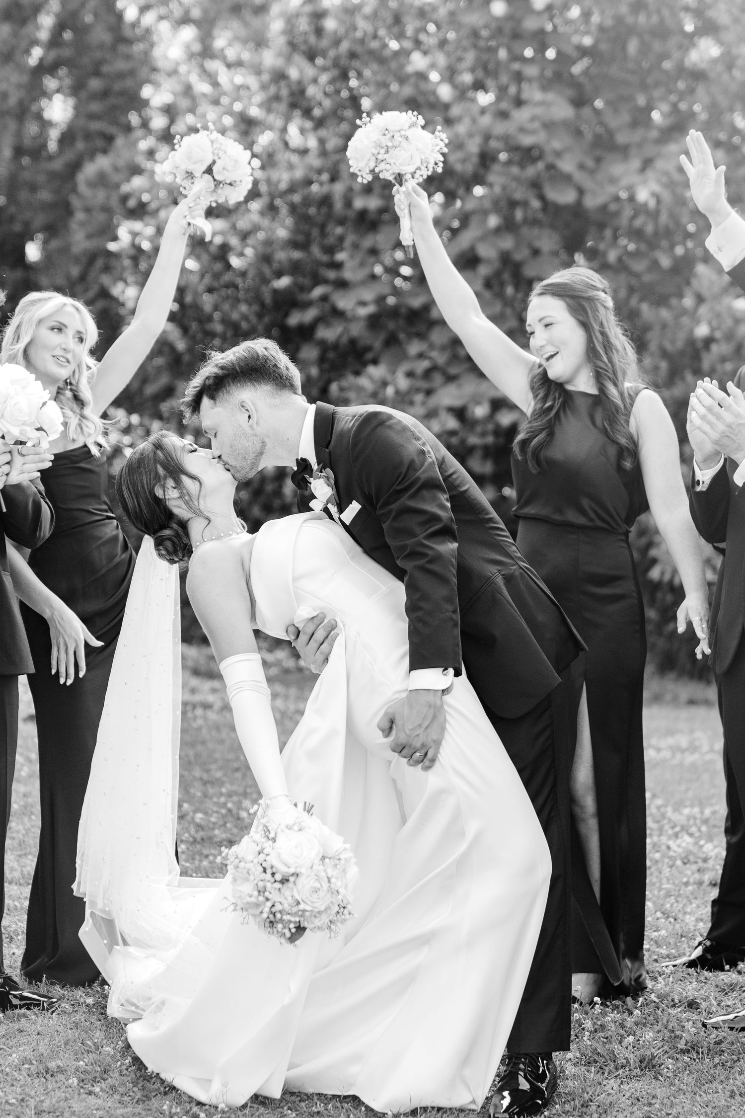 Fun wedding party photo series showing groomsmen in black suits dipping bridesmaids in white dresses outdoors.