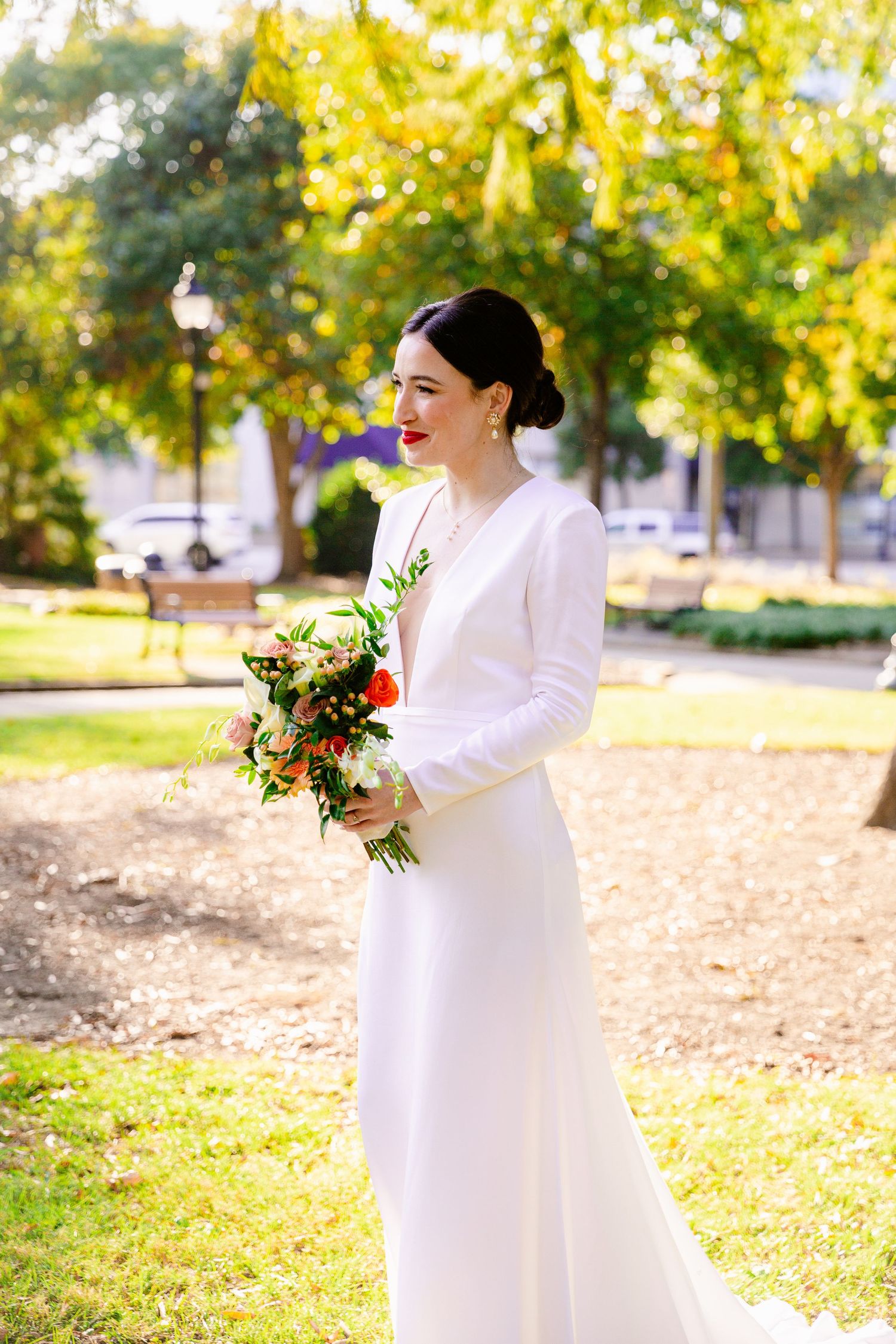 A bride in an elegant white long-sleeved gown holds a colorful bouquet in a sunny park.