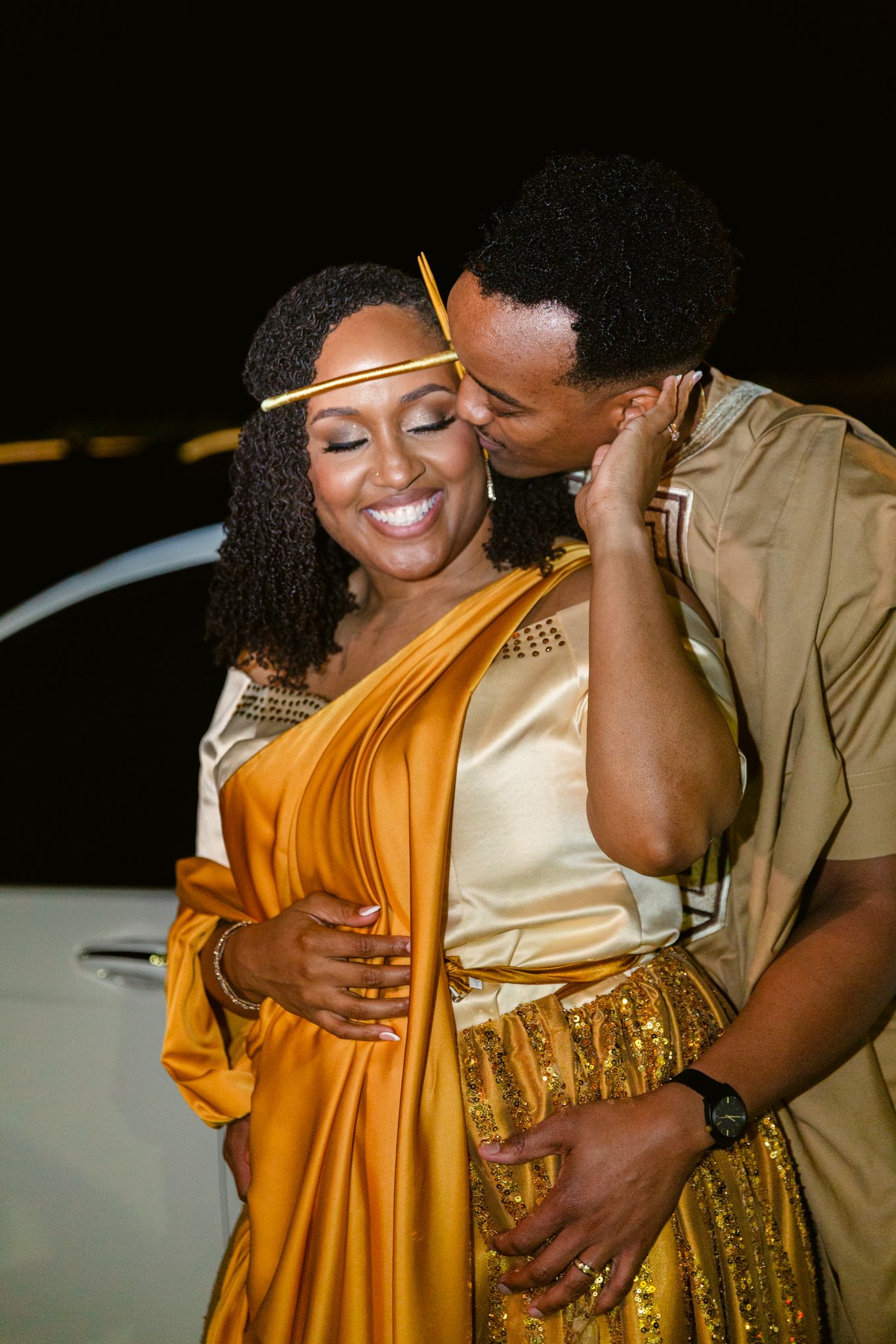 A couple shares an intimate moment in formal attire with gold and mustard yellow colors against a dark background.