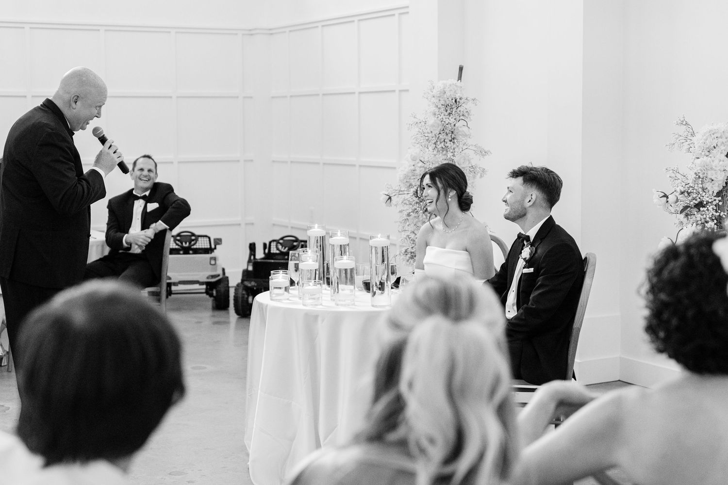 Elegant white wedding reception venue with tables and guests during speeches.
