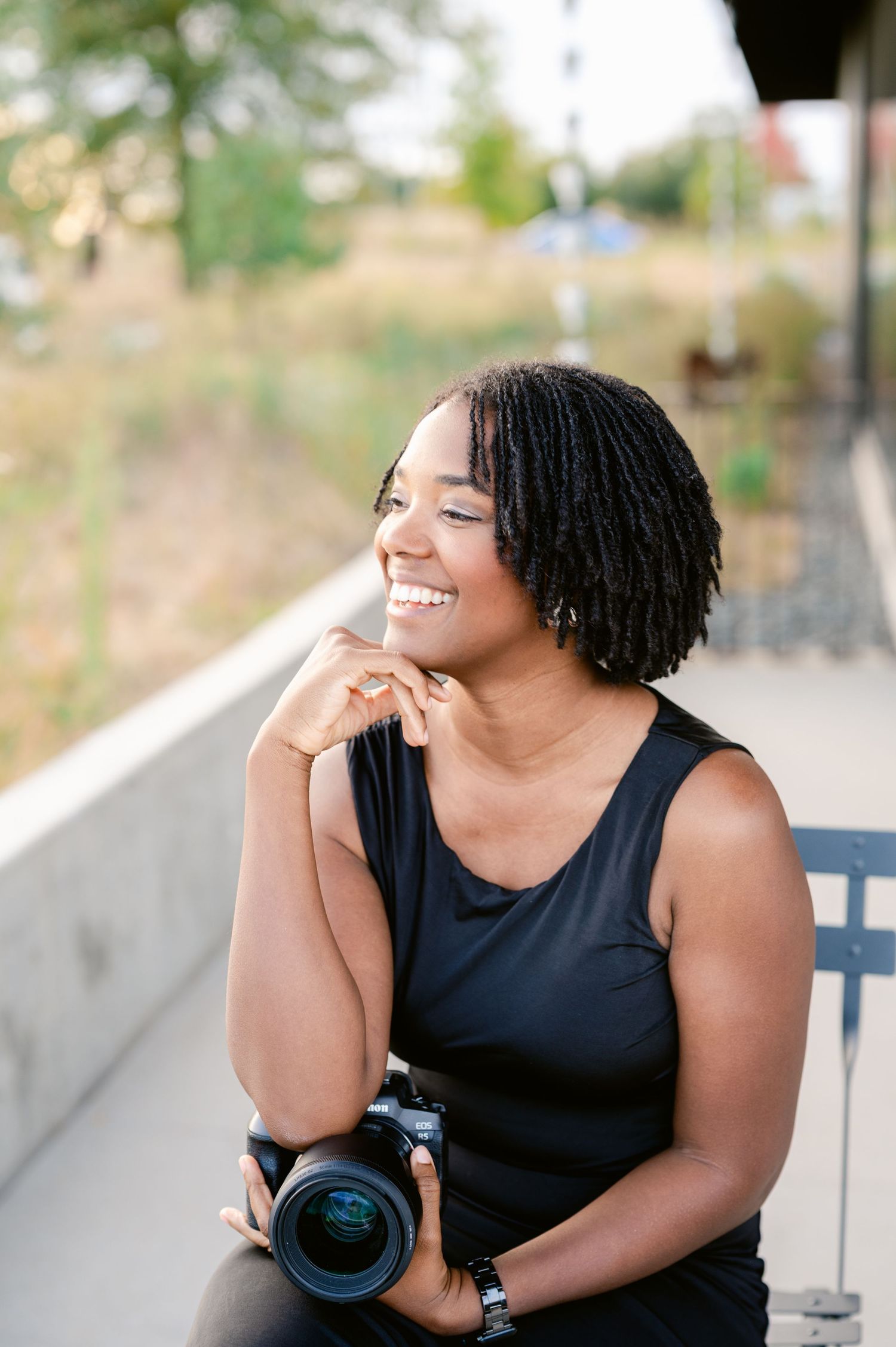 A photographer with natural hair and a black tank top smiles while holding a professional camera outdoors.