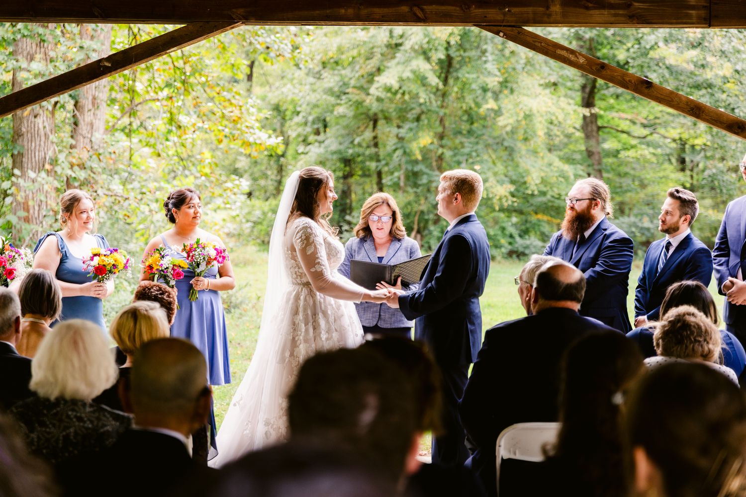 Wedding ceremony guests seated outdoors under a wooden pavilion watching the ceremony proceedings.