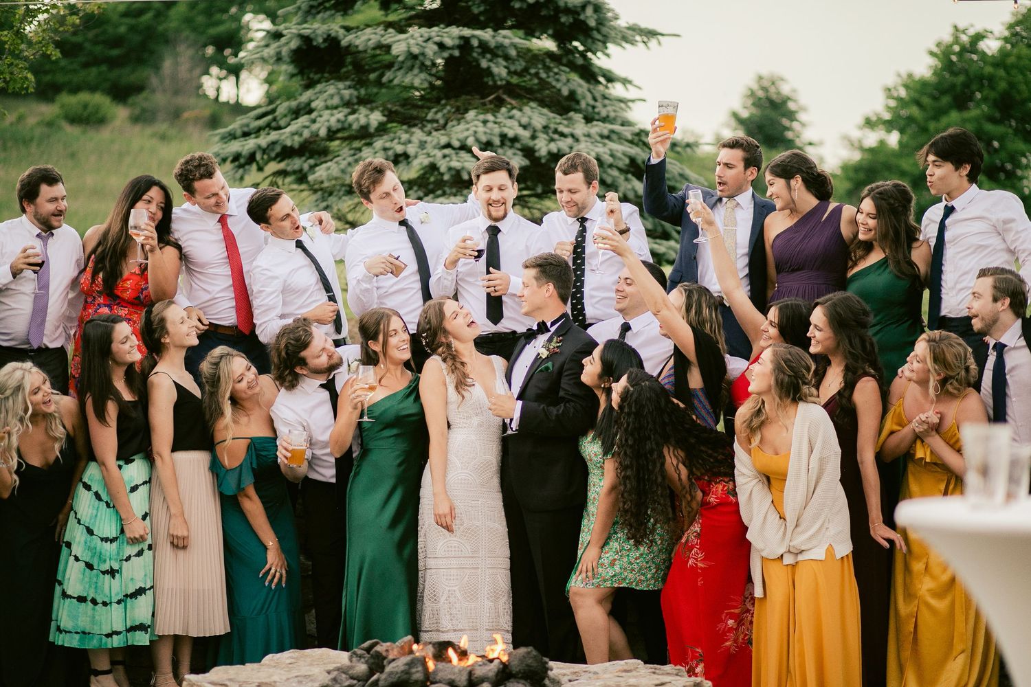 Wedding party lined up outdoors wearing green, pink, and yellow dresses with trees in the background.