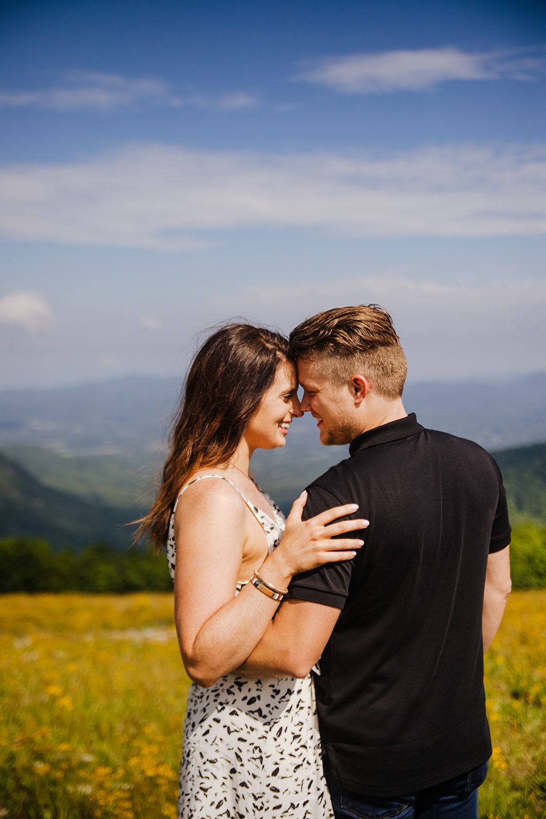 A romantic couple embraces on a mountaintop meadow filled with yellow wildflowers under a blue summer sky.