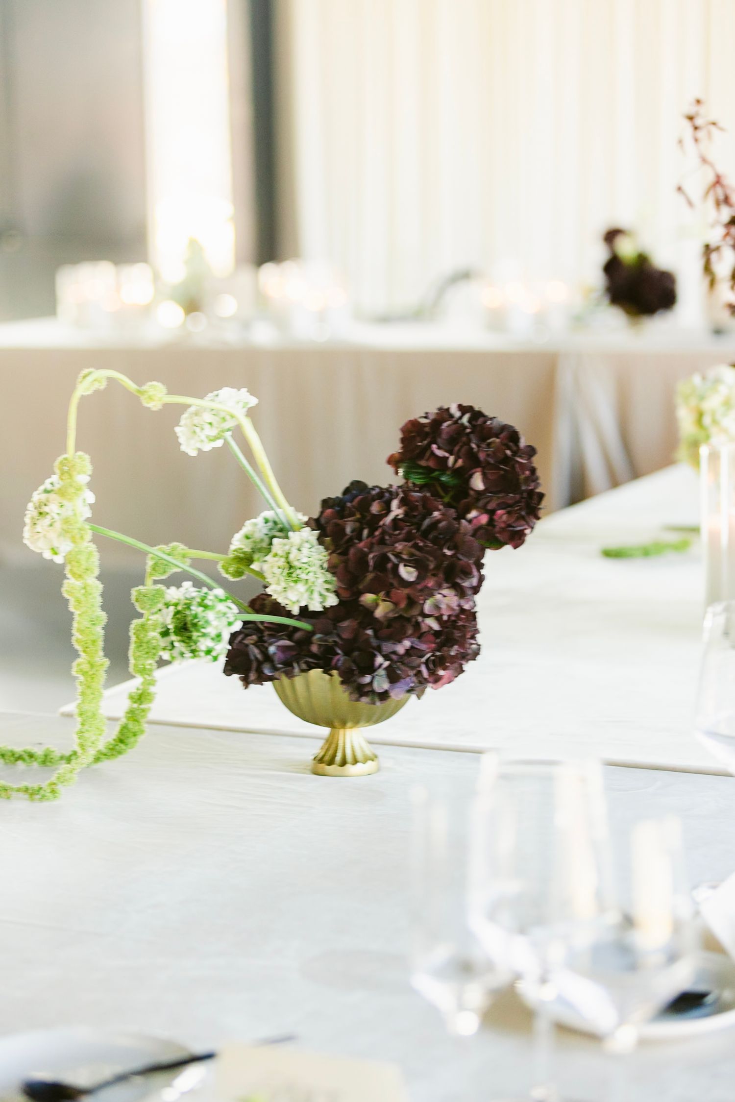 Elegant floral centerpieces with dark purple hydrangeas and green accents arranged on a white tablecloth.