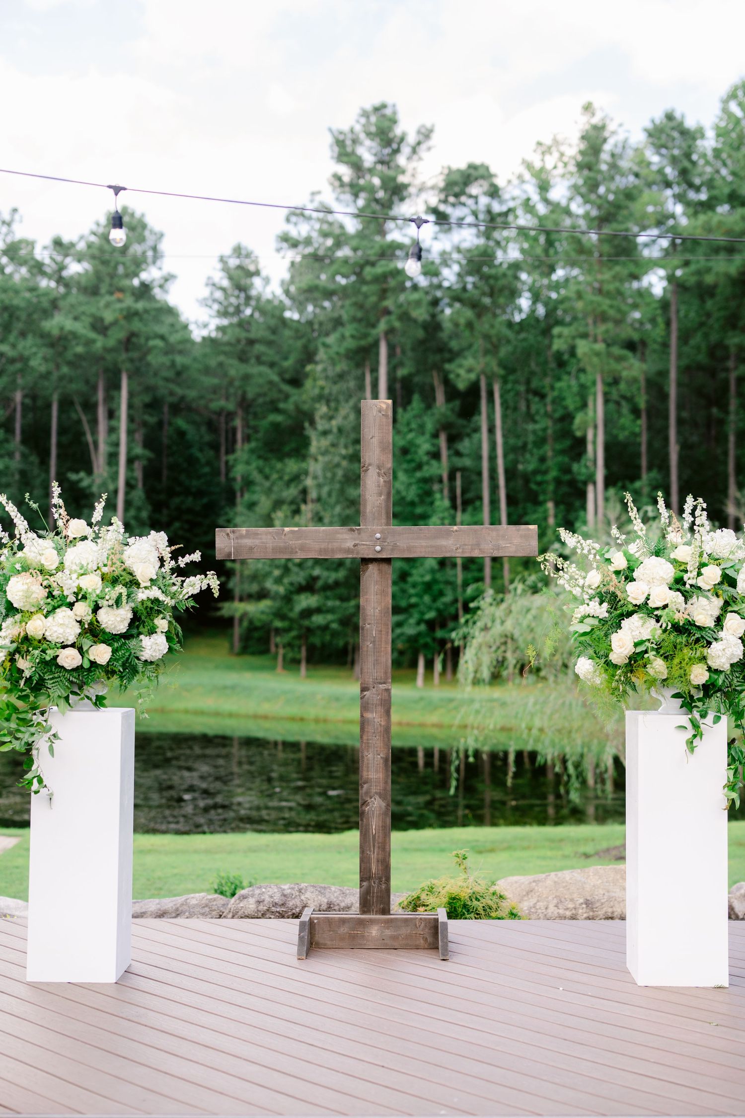 Wooden crosses and white floral arrangements line a rustic outdoor wedding ceremony space with pine trees in background.