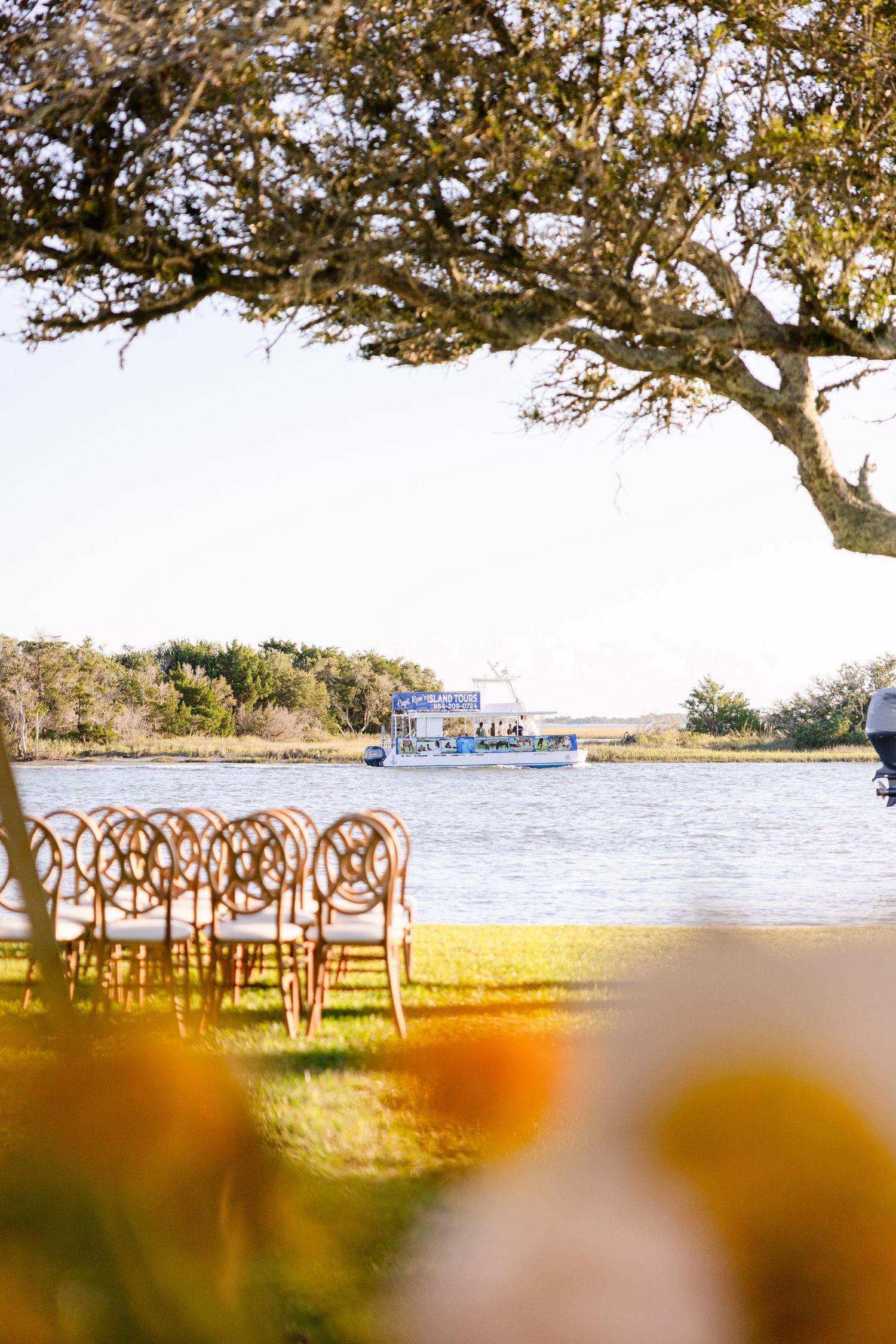Panoramic view of a waterfront wedding venue with wooden chairs arranged under a tree by the ocean.