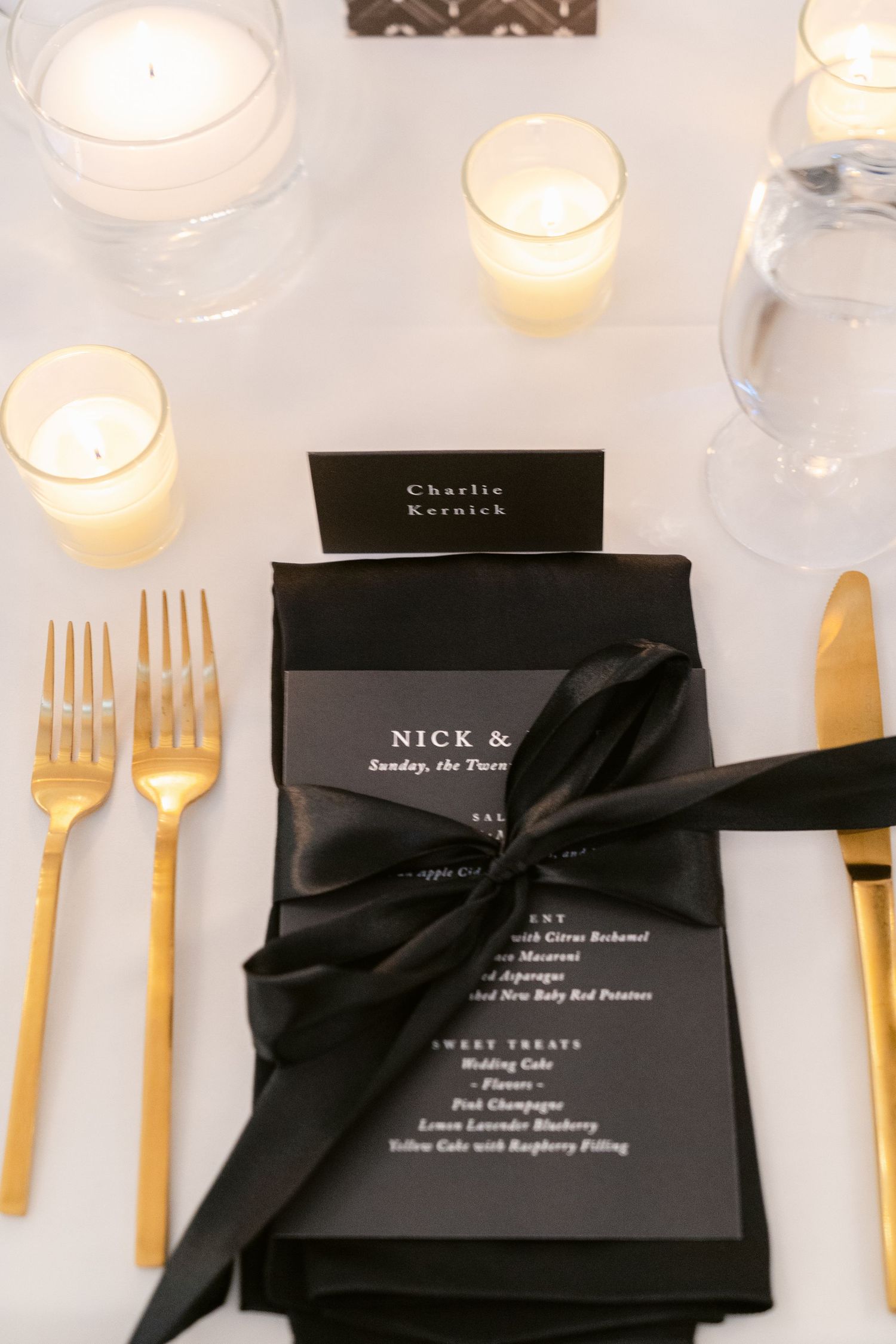 Elegant black wedding menus with gold utensils and glowing white candles arranged on a formal dining table.