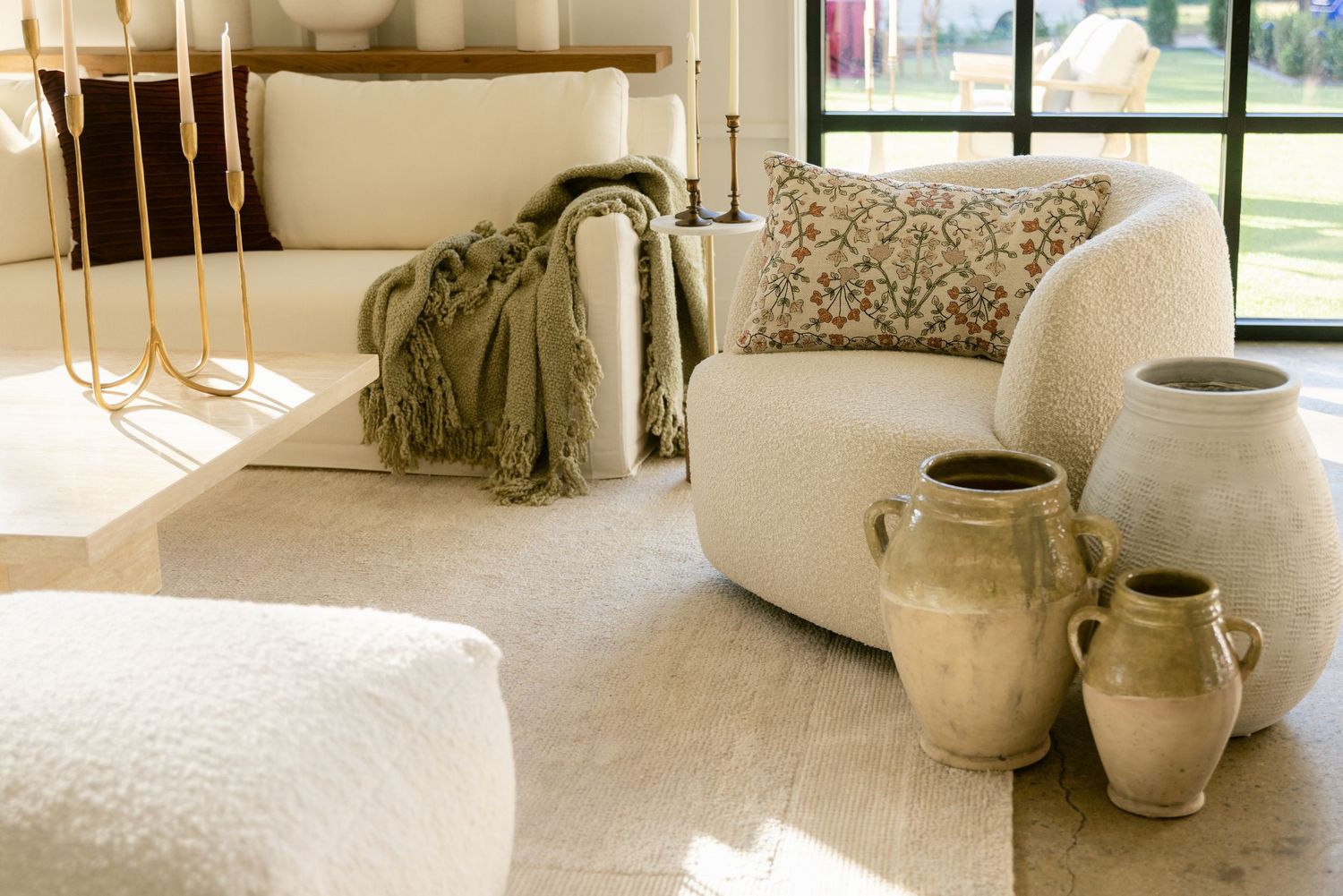 Cozy living room corner with cream curved chair, olive throw blanket, and rustic ceramic vases on hardwood floor.