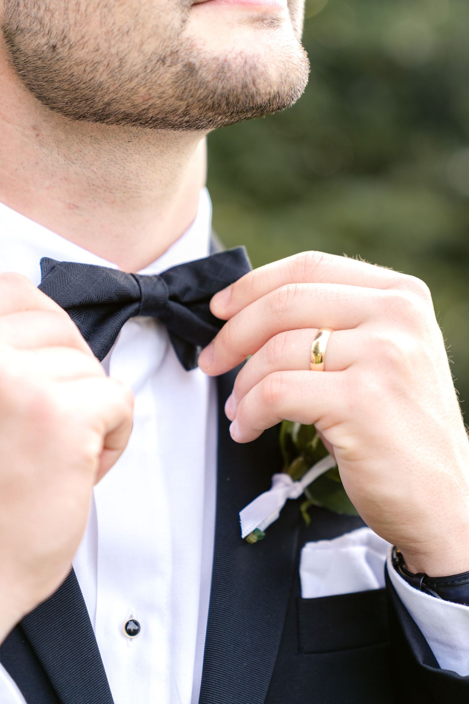 Close-up of a groom adjusting his black bow tie and showing gold wedding band against formal attire.