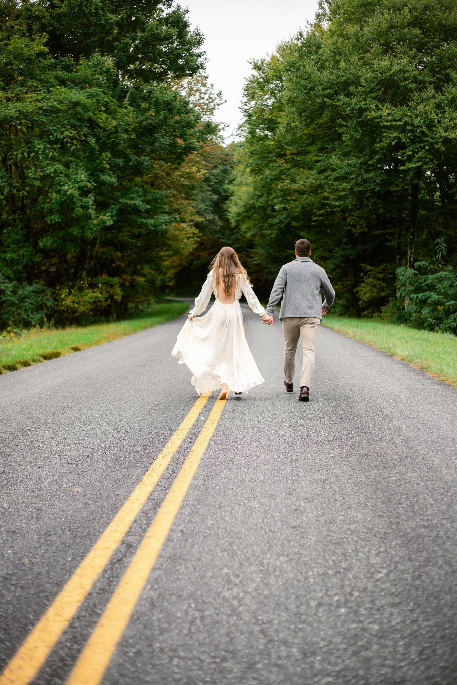 Couple walking down a tree-lined country road holding hands with the bride wearing a flowing white dress.