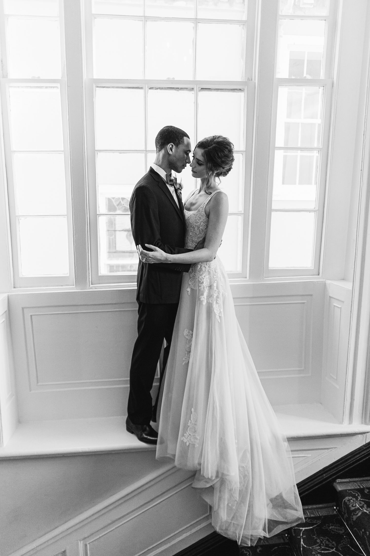 A romantic black and white wedding photo of a couple embracing by large windows in an elegant room.