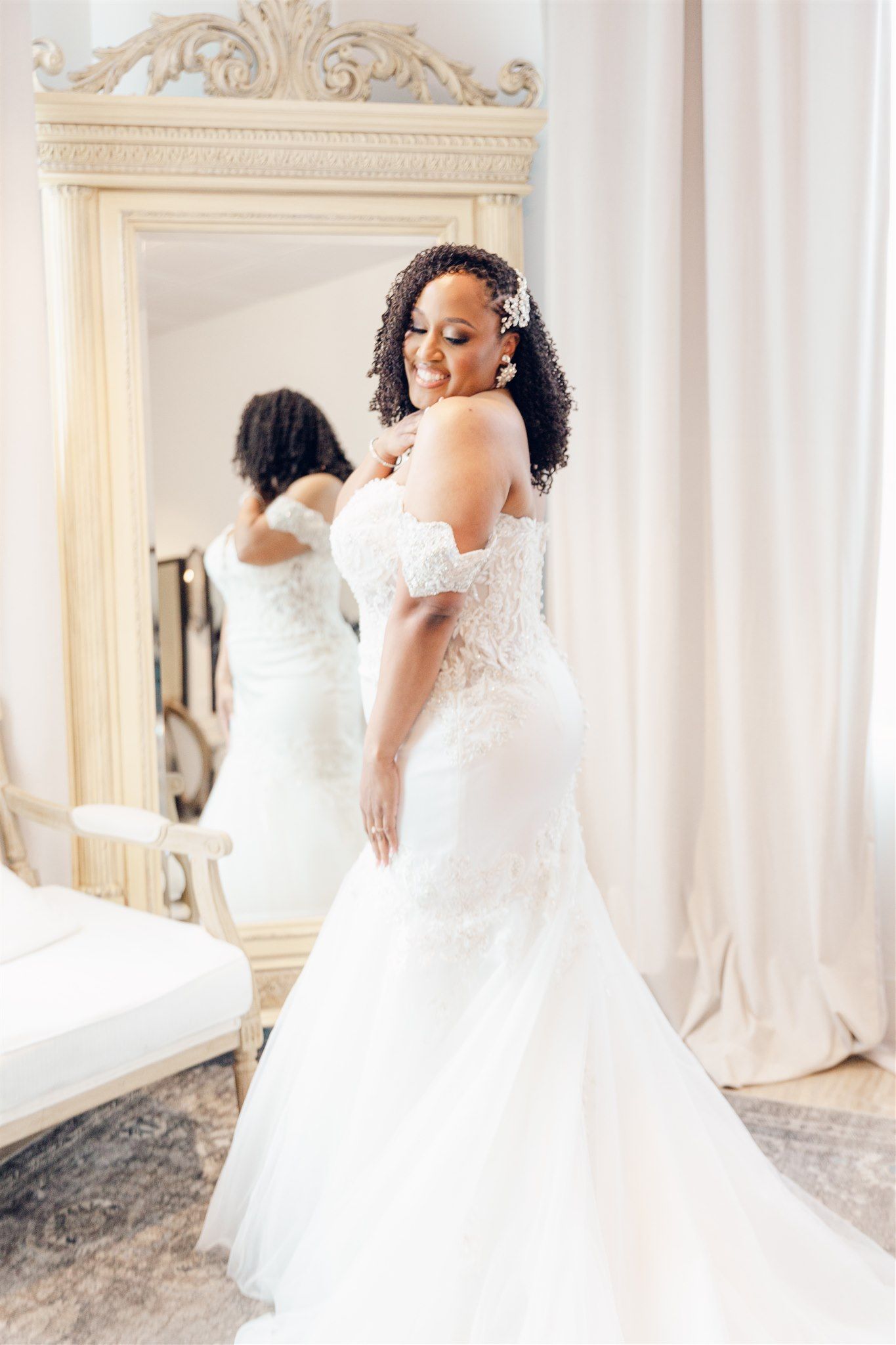 A bride in a flowing white off-shoulder wedding gown twirls gracefully in an elegant room with ornate mirrors.