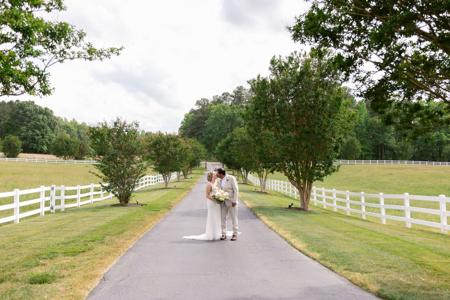 Couple walking along a scenic country road lined with white fences and green trees on a sunny summer day.