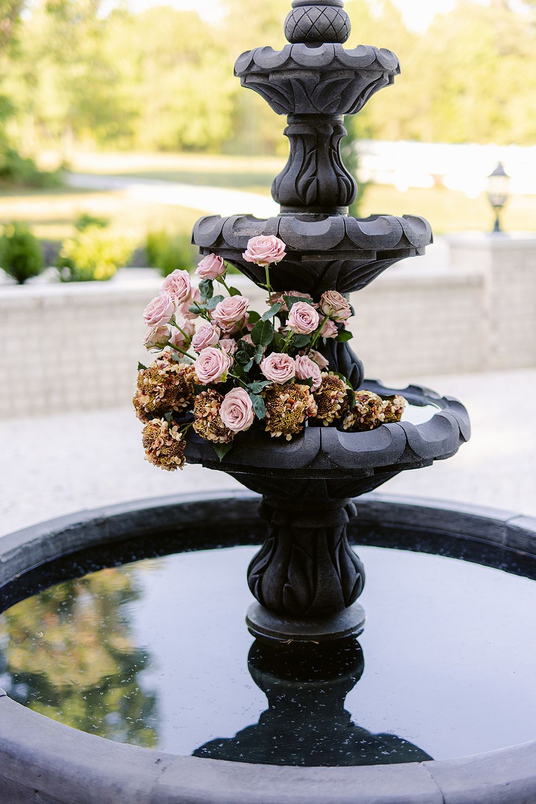 Black ornate fountain decorated with pink roses and dried hydrangeas in an outdoor garden setting.