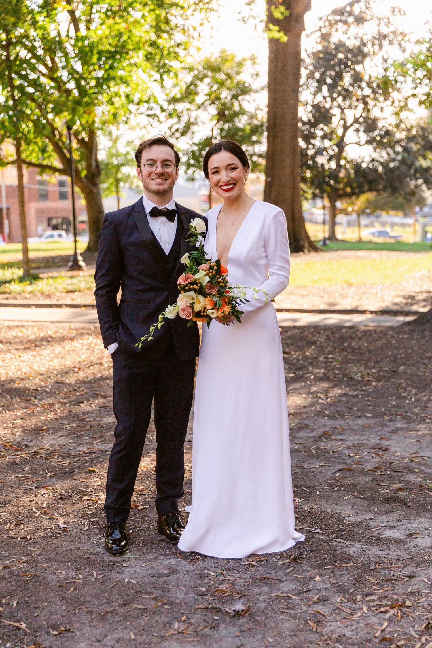 A wedding couple in formal attire poses playfully outdoors on fallen autumn leaves in a park setting.