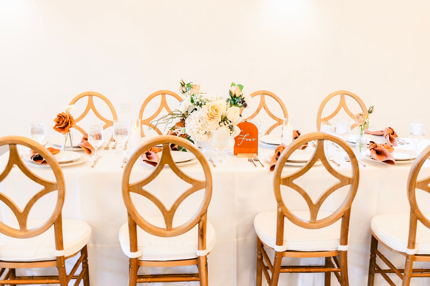 White wooden cross-back dining chairs lined up in a row against a light beige wall at an elegant event venue.
