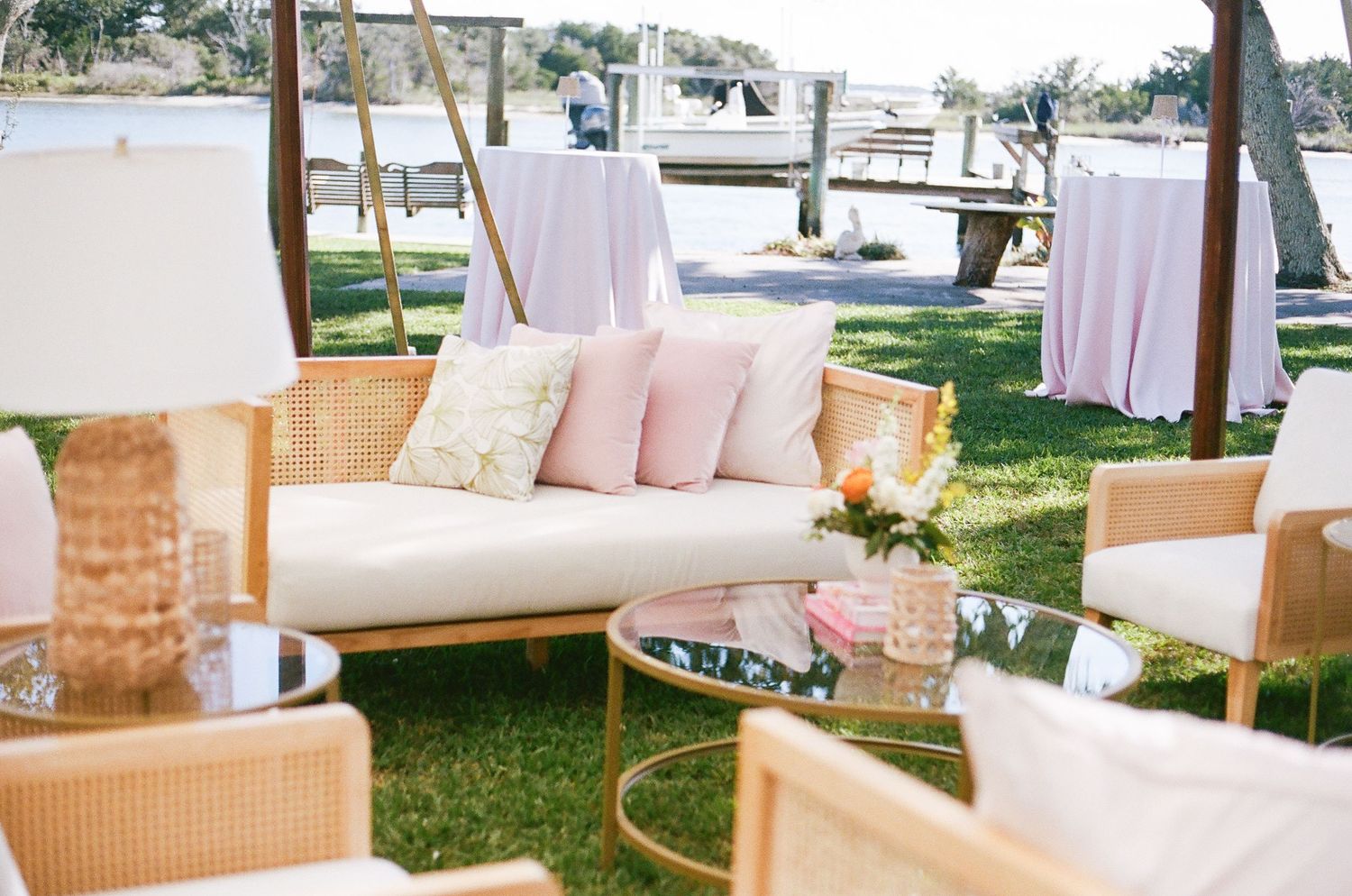 Elegant outdoor lounge area with white sofa, blush pillows and rattan furniture on a grassy lawn.