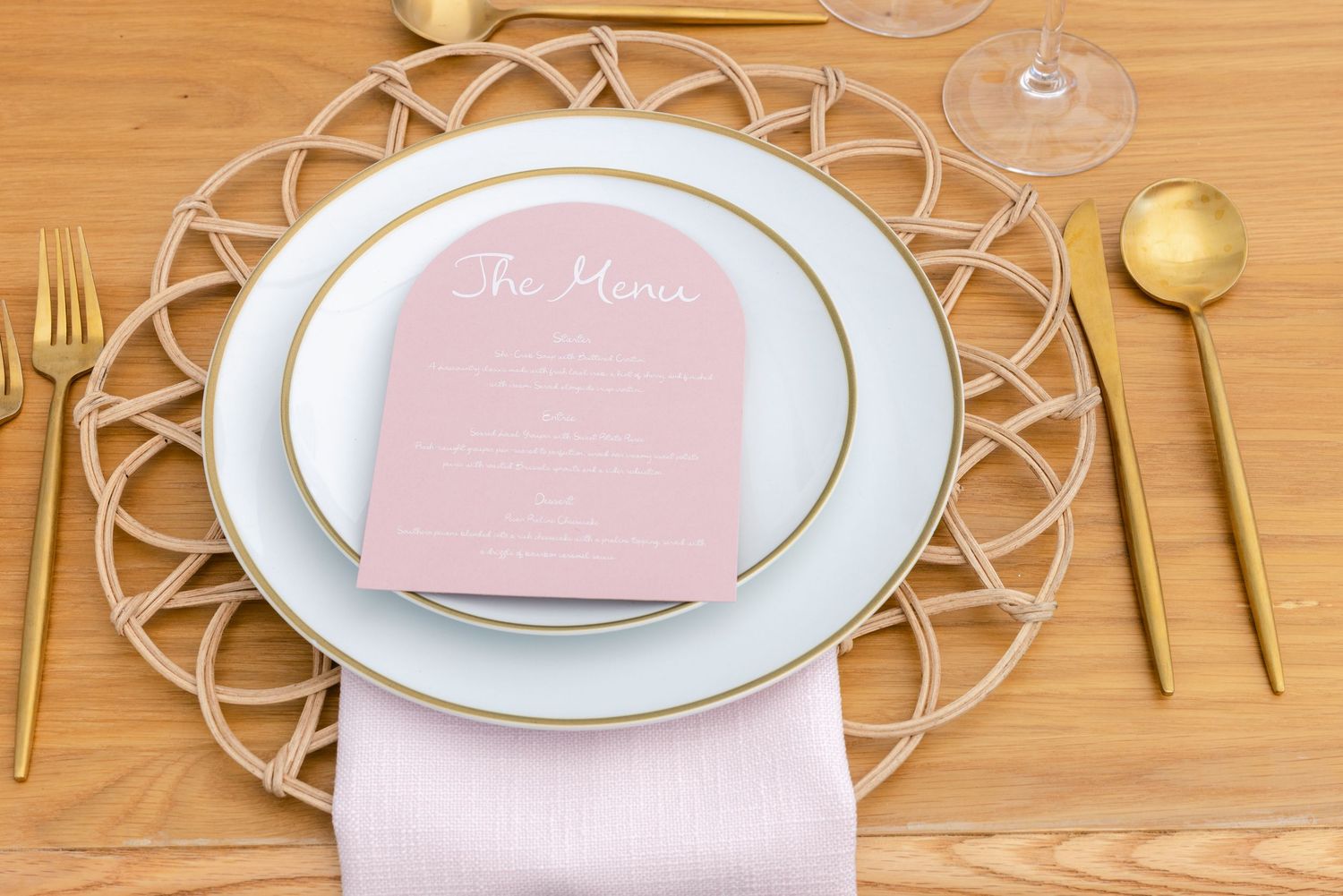 Elegant place setting with pink menu card, gold flatware, and white charger plate on wooden table.