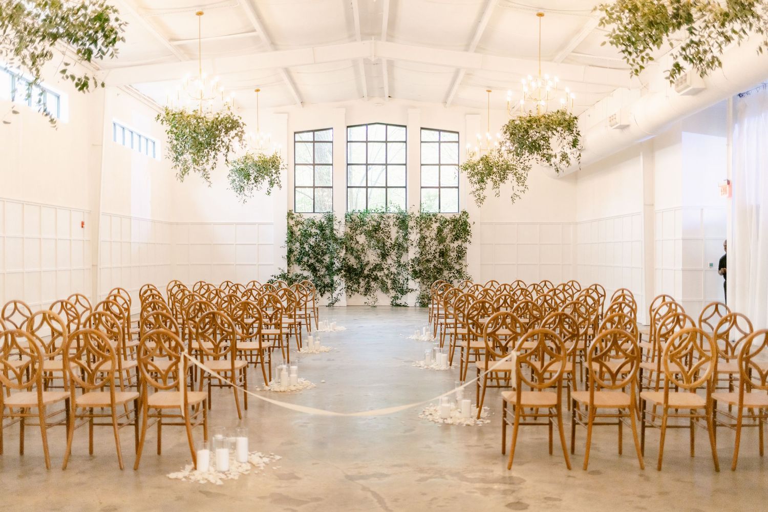 Light-filled white event space with rows of wooden chairs arranged for a ceremony with sunlight streaming through windows.