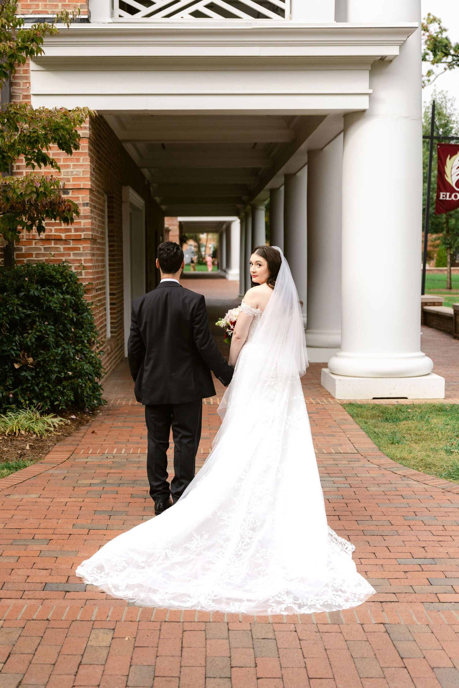 A wedding couple walks along a columned brick pathway with a flowing white lace train trailing behind.