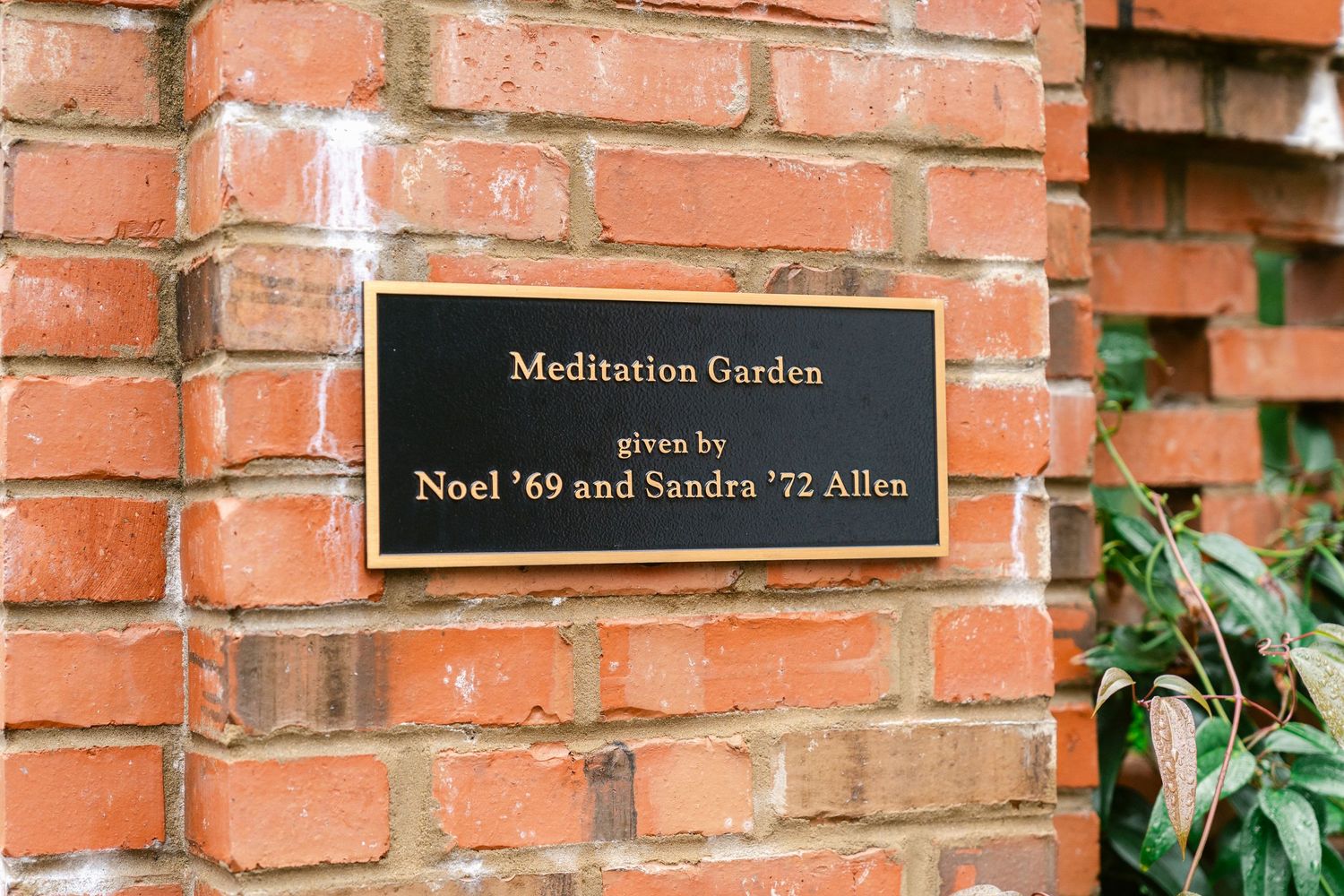 A row of black name plaques mounted on a red brick wall with green foliage visible.