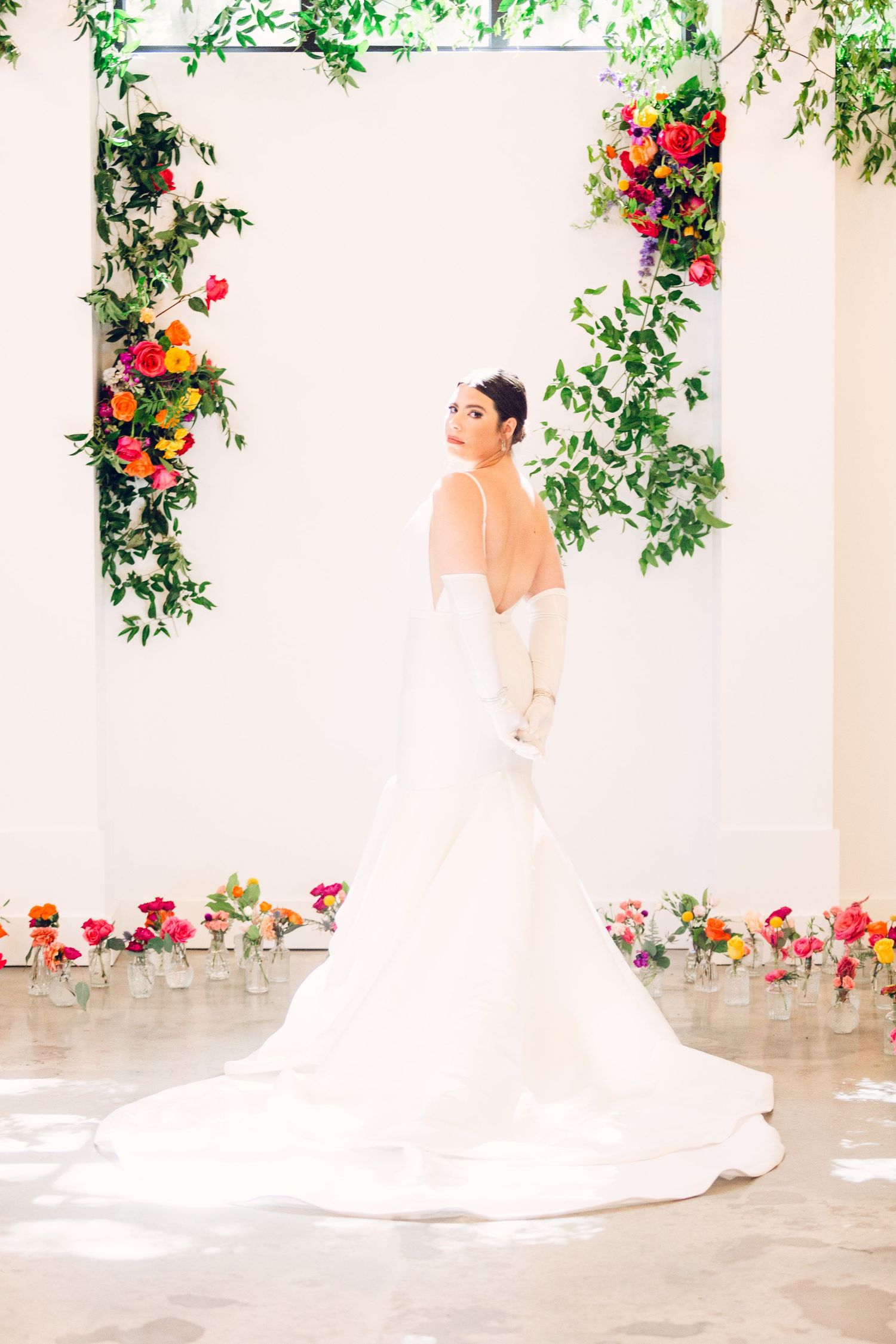 Elegant bride in flowing white gown poses under archway decorated with vibrant red and yellow floral arrangements.
